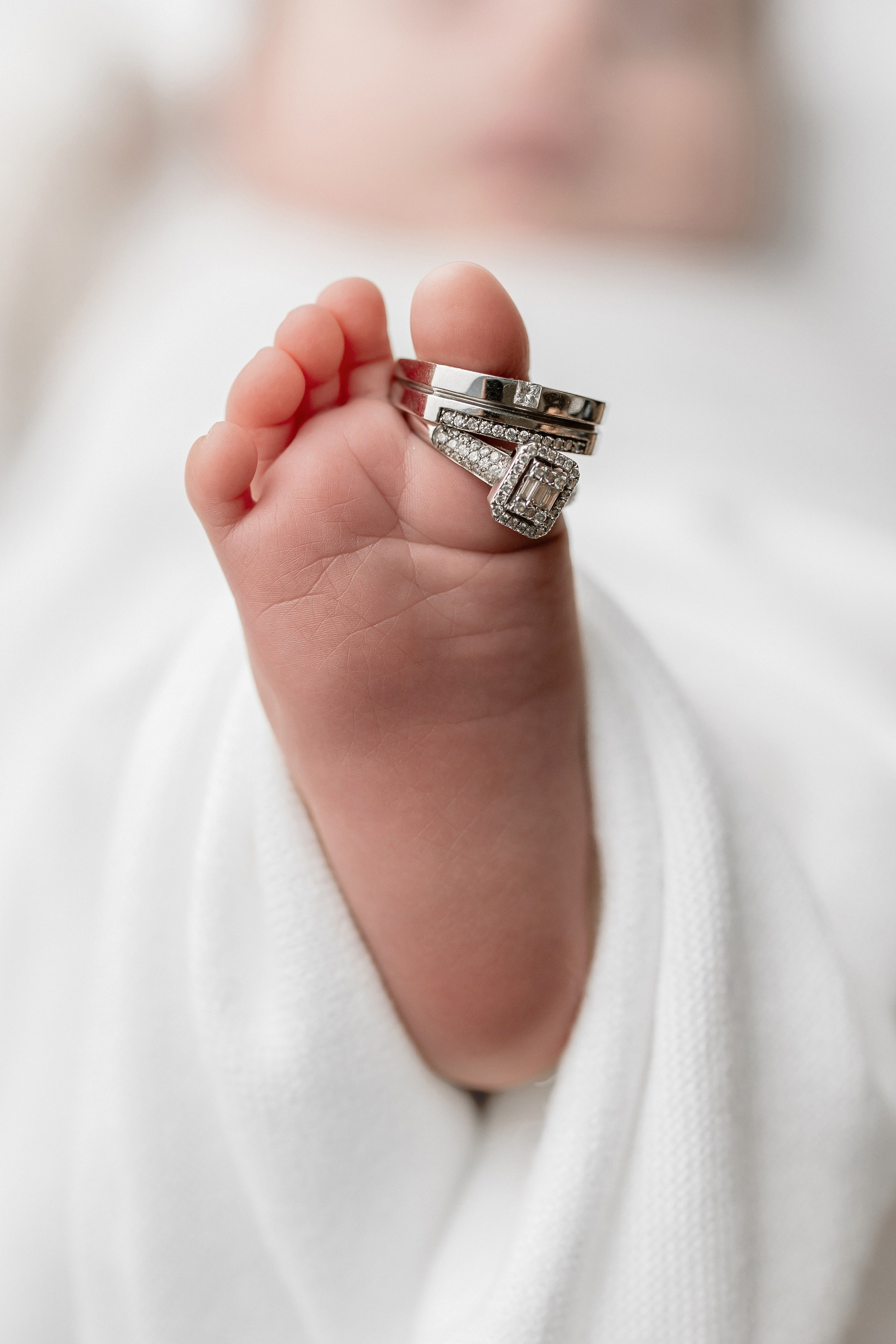 Close-up of newborn baby foot wrapped in white blanket with wedding rings on toes — symbolic newborn photography capturing love and family bonds.