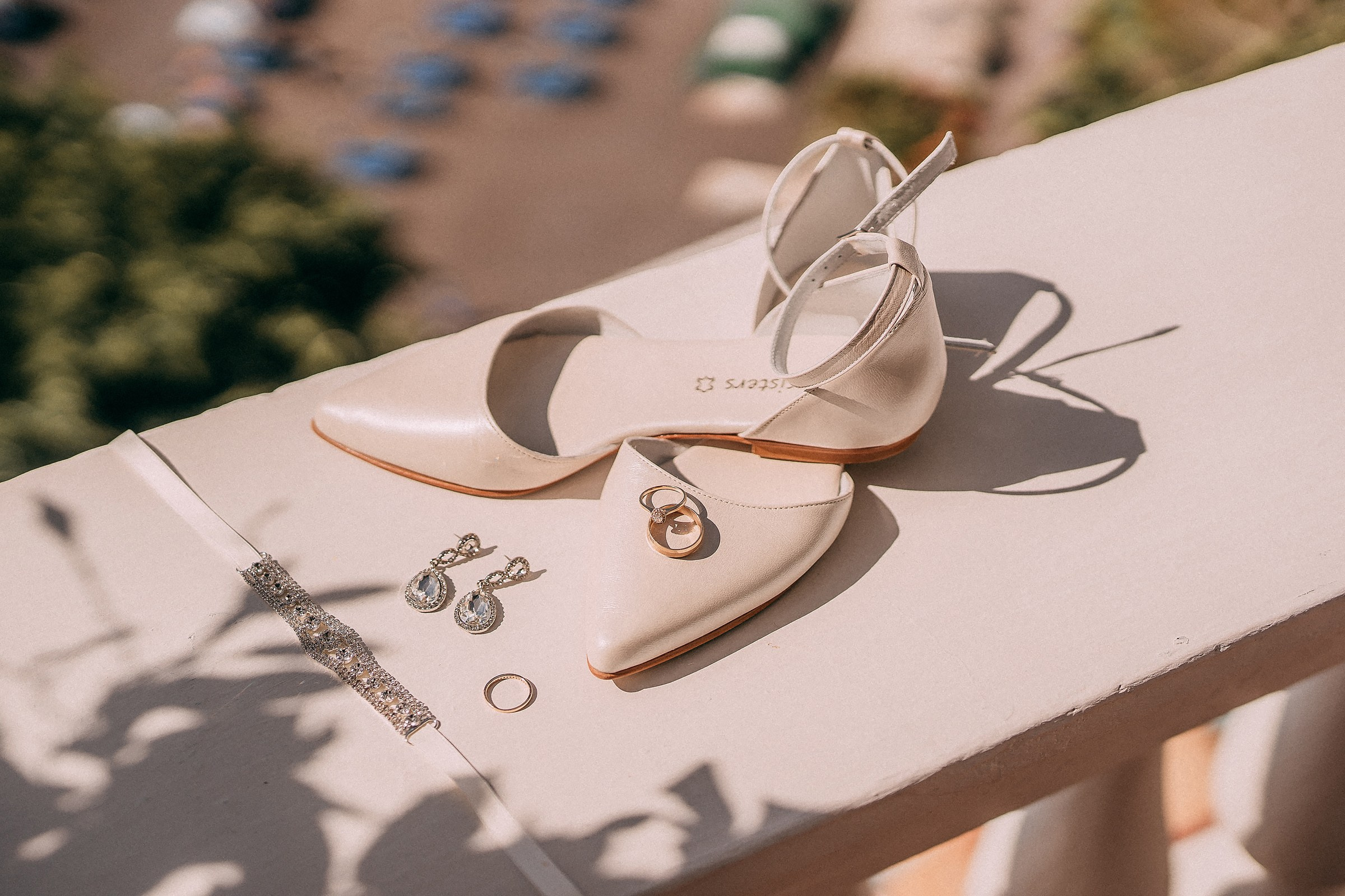 A minimalist wedding accessory setup showcasing ivory bridal shoes, earrings, a bracelet, and wedding rings arranged on a sunlit terrace overlooking Positano’s coastal beauty.