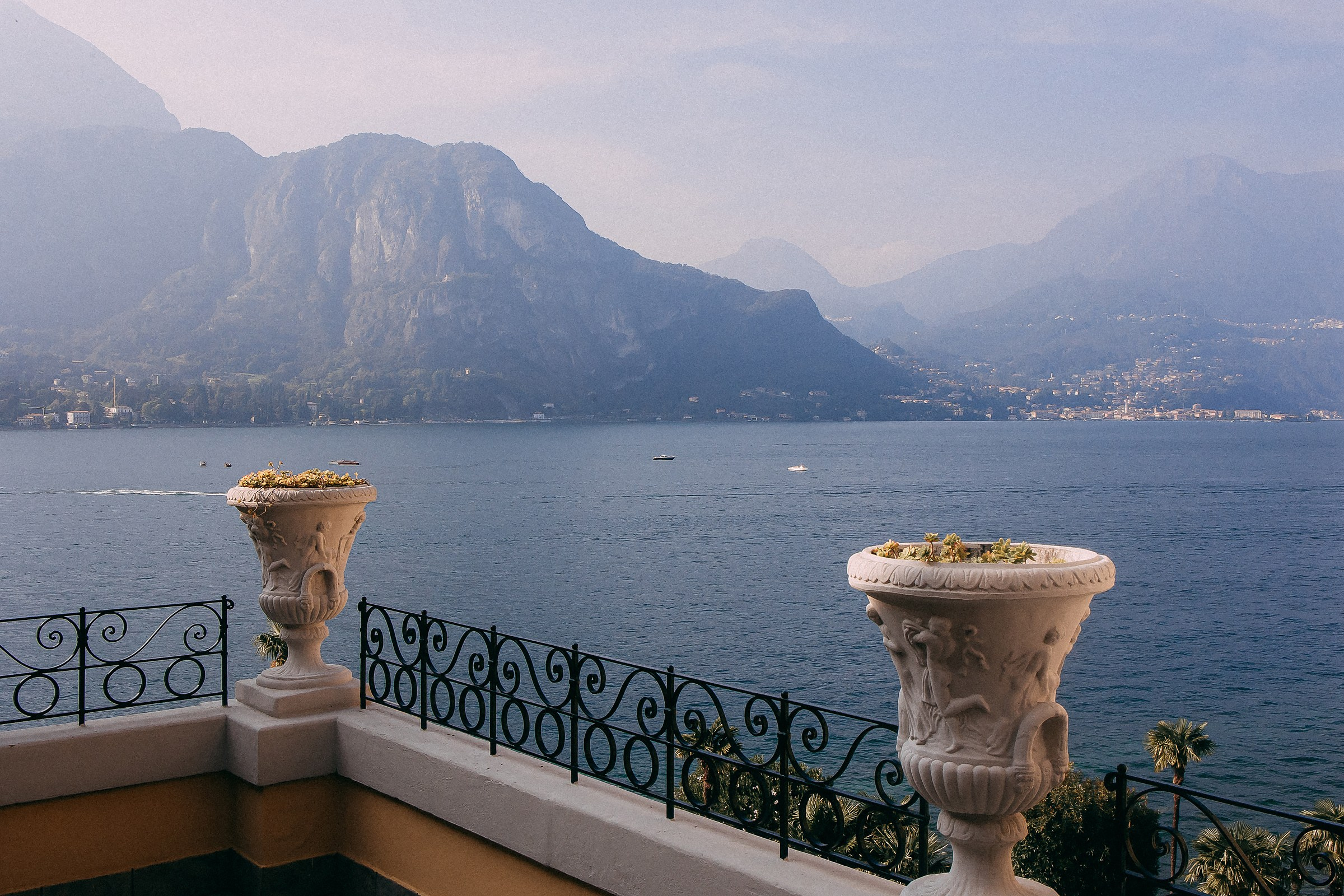 A breathtaking view of a serene lake Como surrounded by towering mountains, framed by an elegant terrace and classical urn planters.