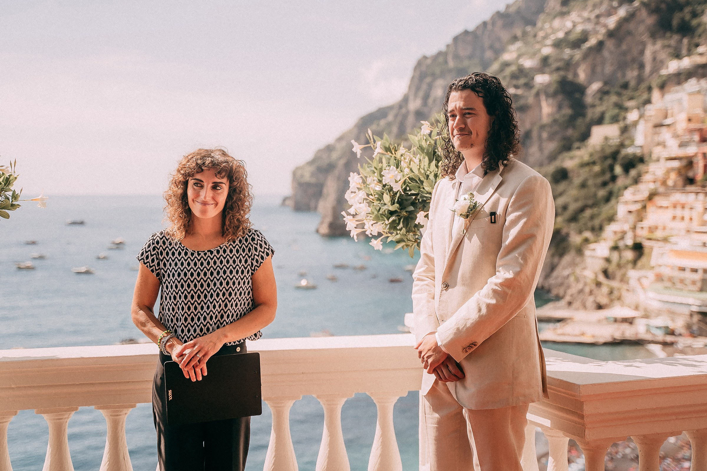 The groom stands with an officiant on a scenic balcony of Villa Boheme, overlooking the sparkling blue sea and vibrant cliffs of Positano under the morning sun.