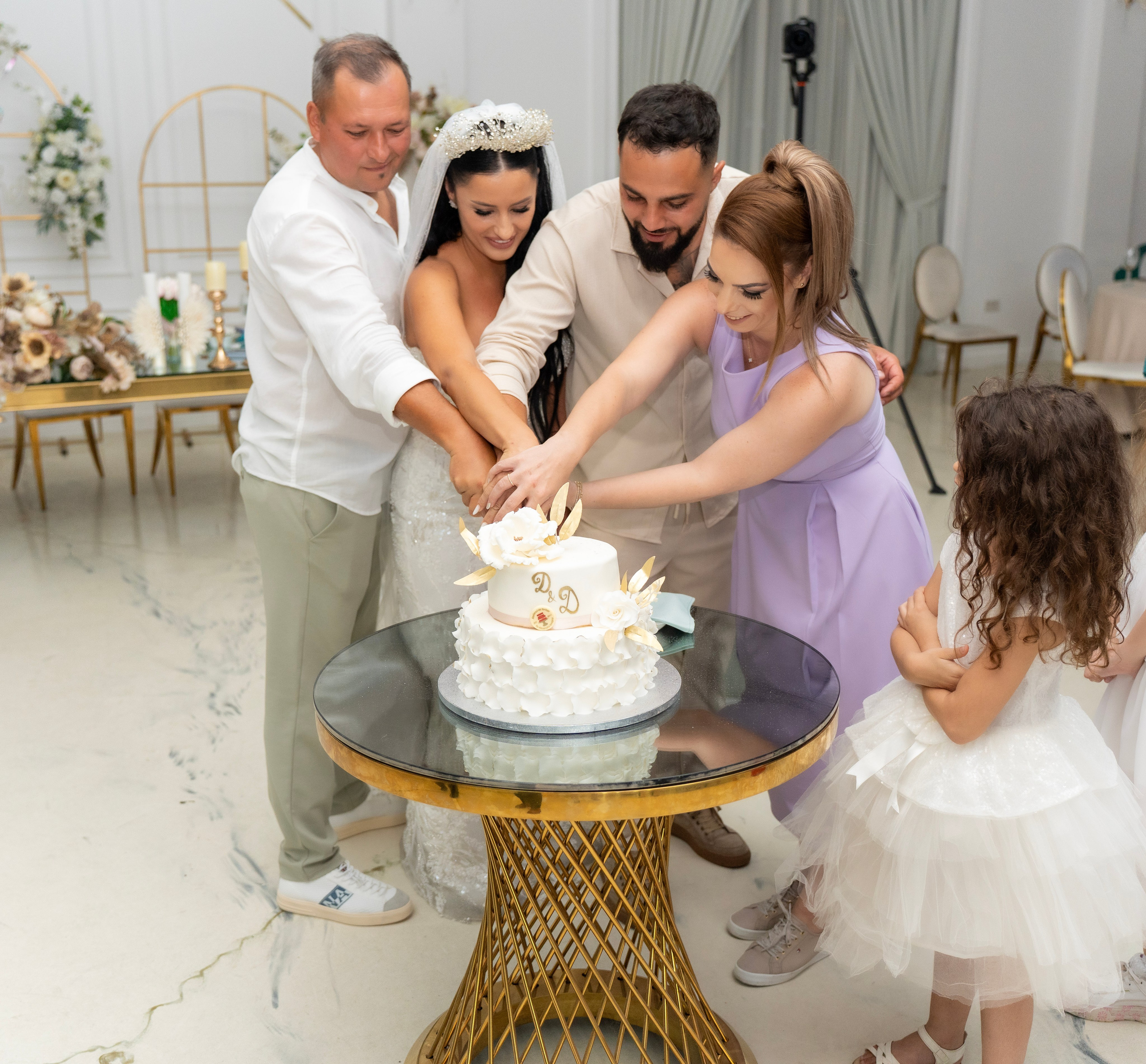 Bride, groom, and two guests cutting a white wedding cake on a glass table at a wedding reception.