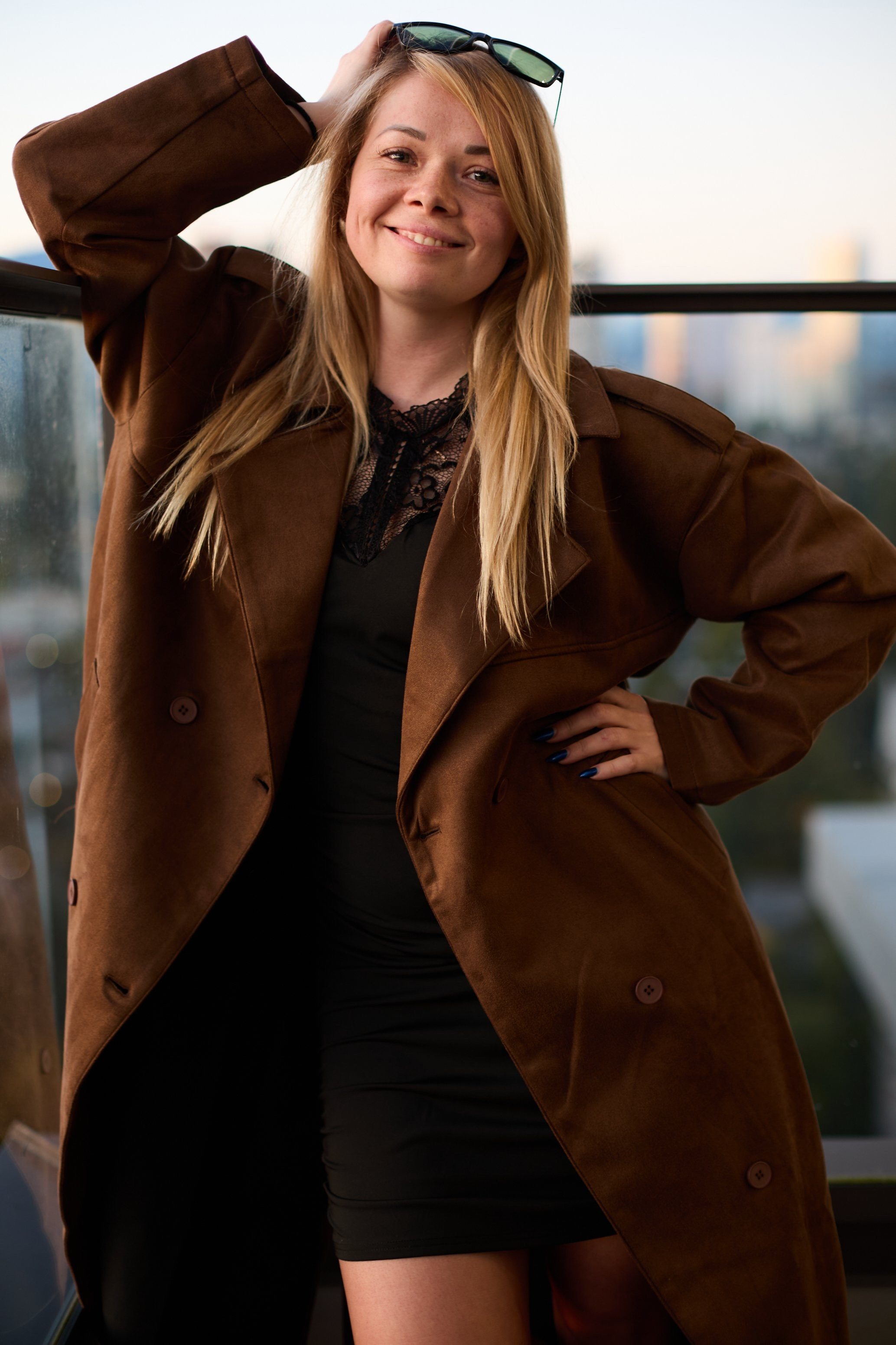 Smiling blonde woman in a long brown coat with sunglasses on her head, posing on a rooftop balcony in Coquitlam