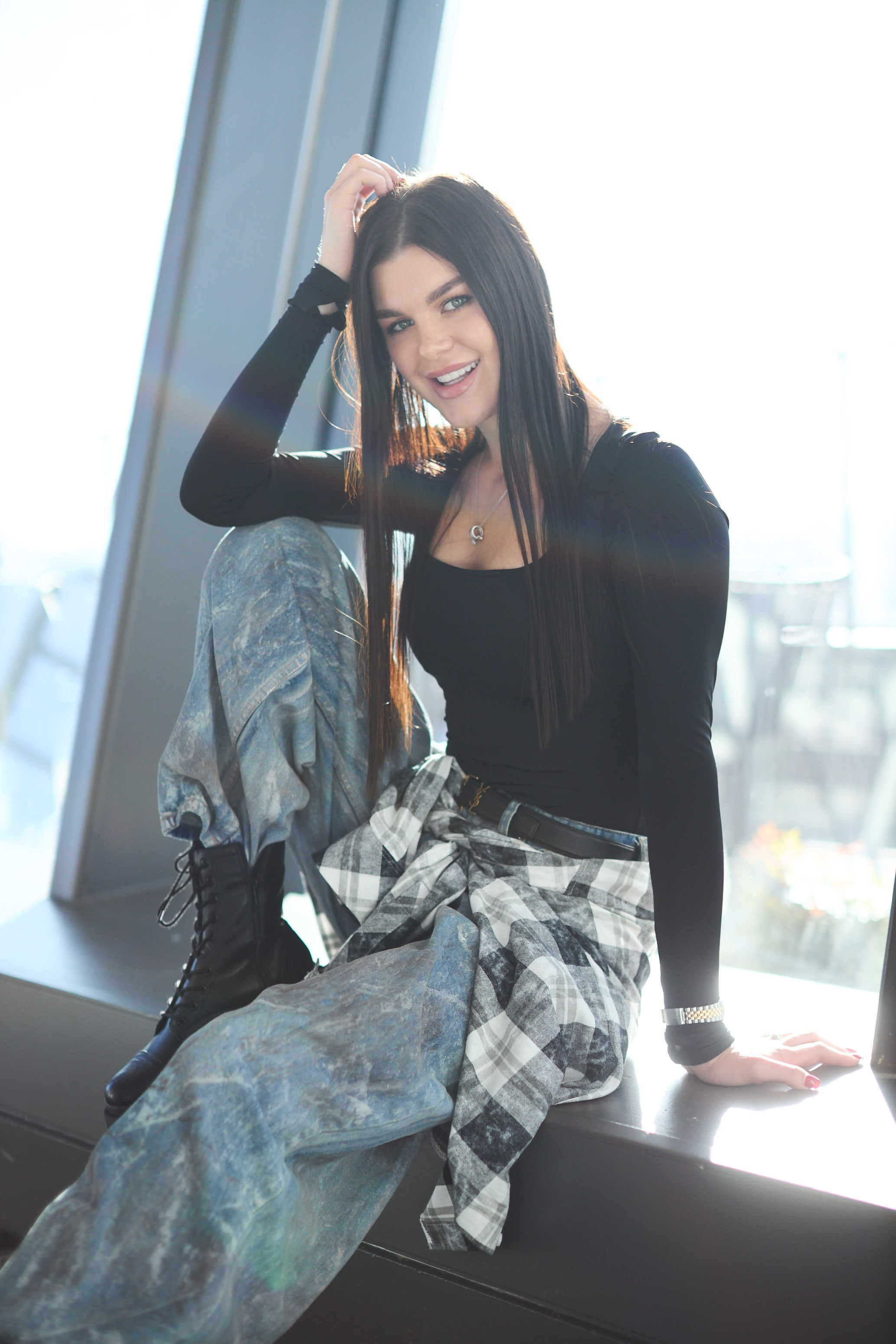 Smiling woman in a black top and grey patterned skirt seated by a floor-to-ceiling window with city views, Coquitlam