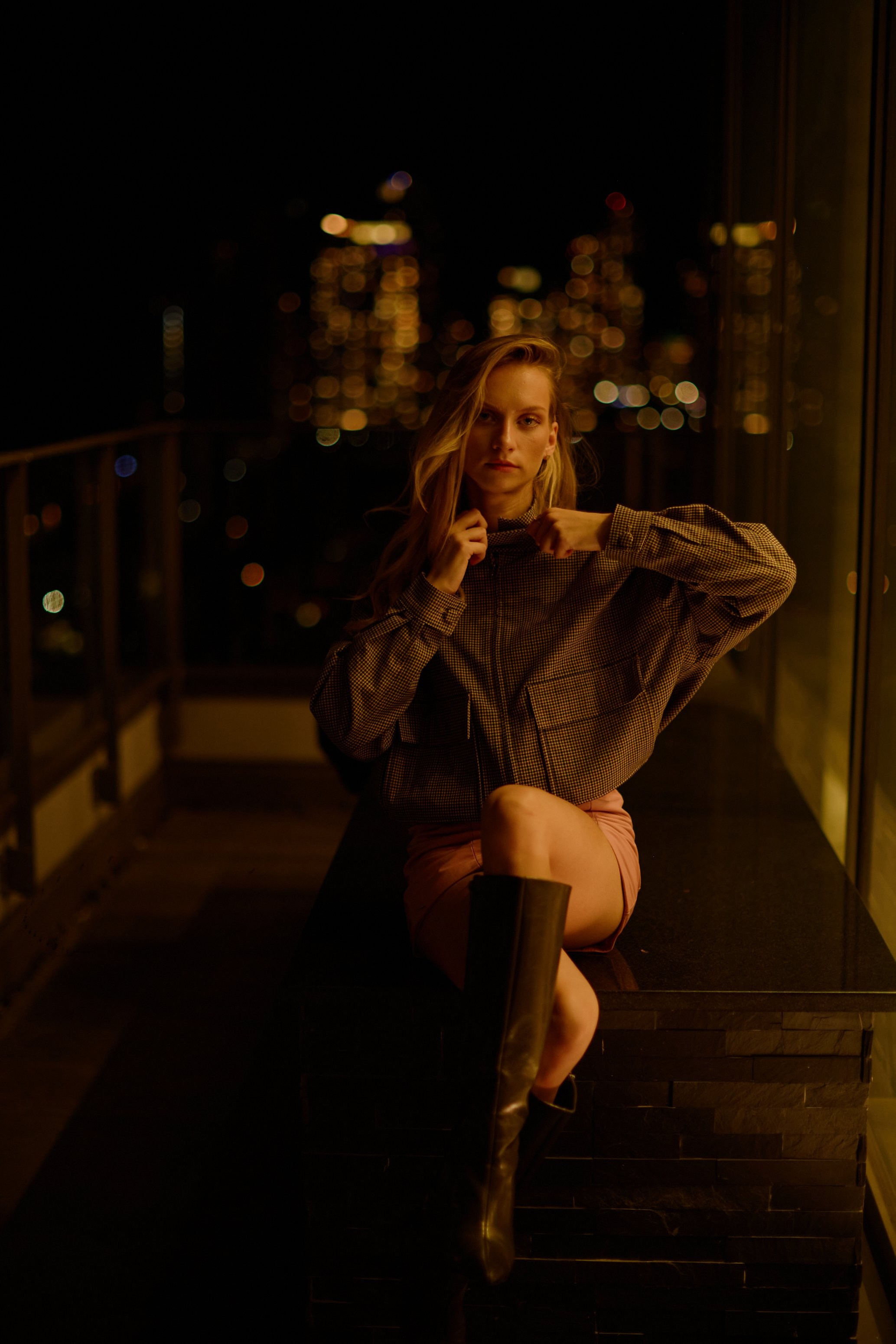 Woman in a brown coat seated in low evening light with city lights behind her — night portrait in Coquitlam