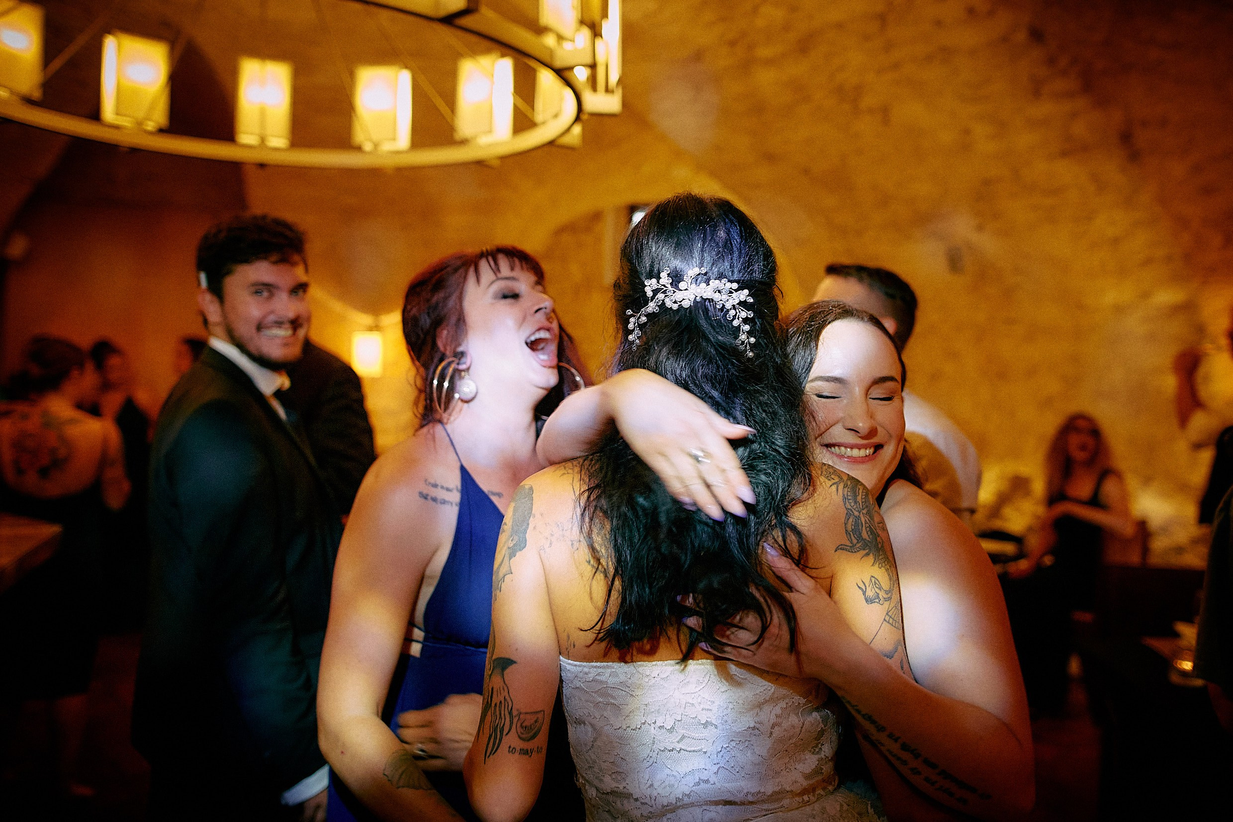 Friends hugging joyful tattooed bride during lively reception.