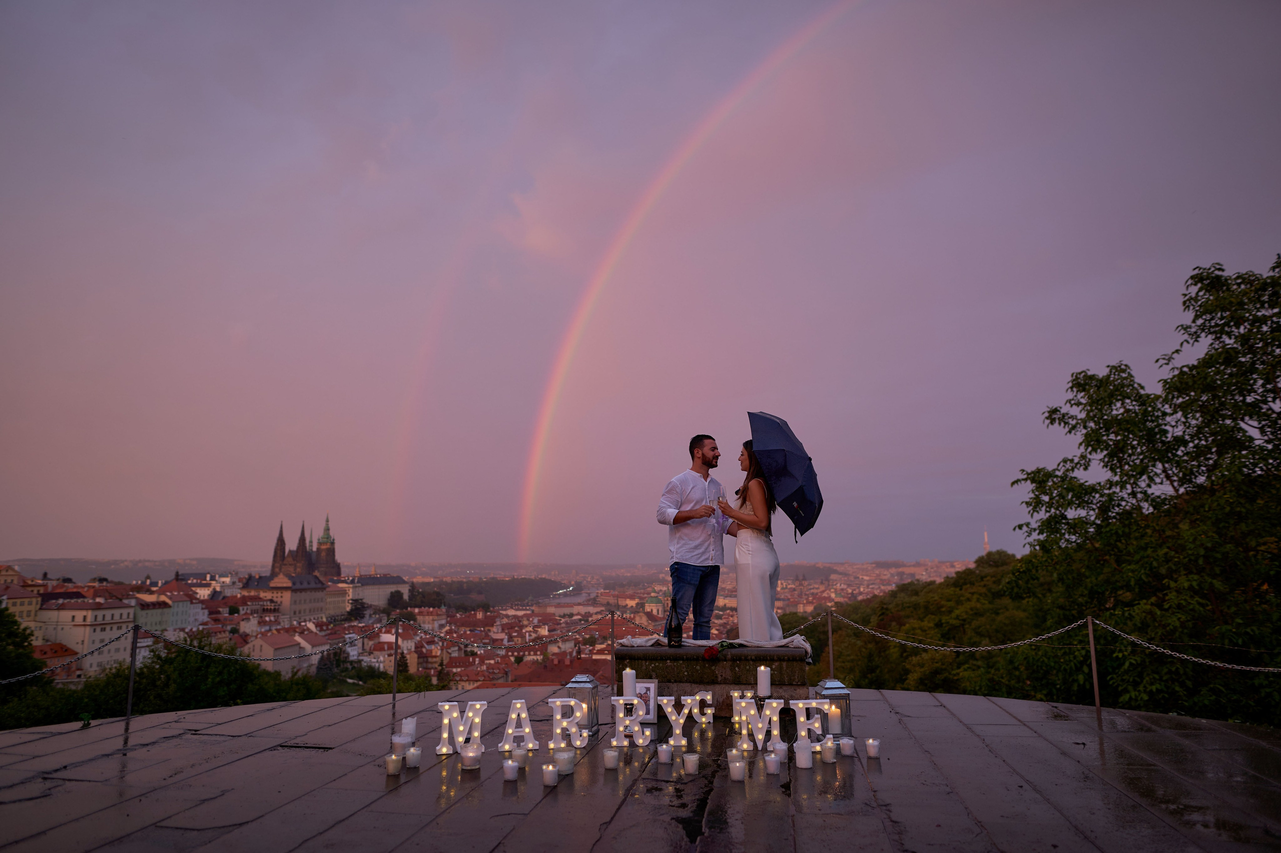 A woman holding an umbrella is embraced by her partner high above Prague, as a purplish-blue sky is filled with two rainbows.