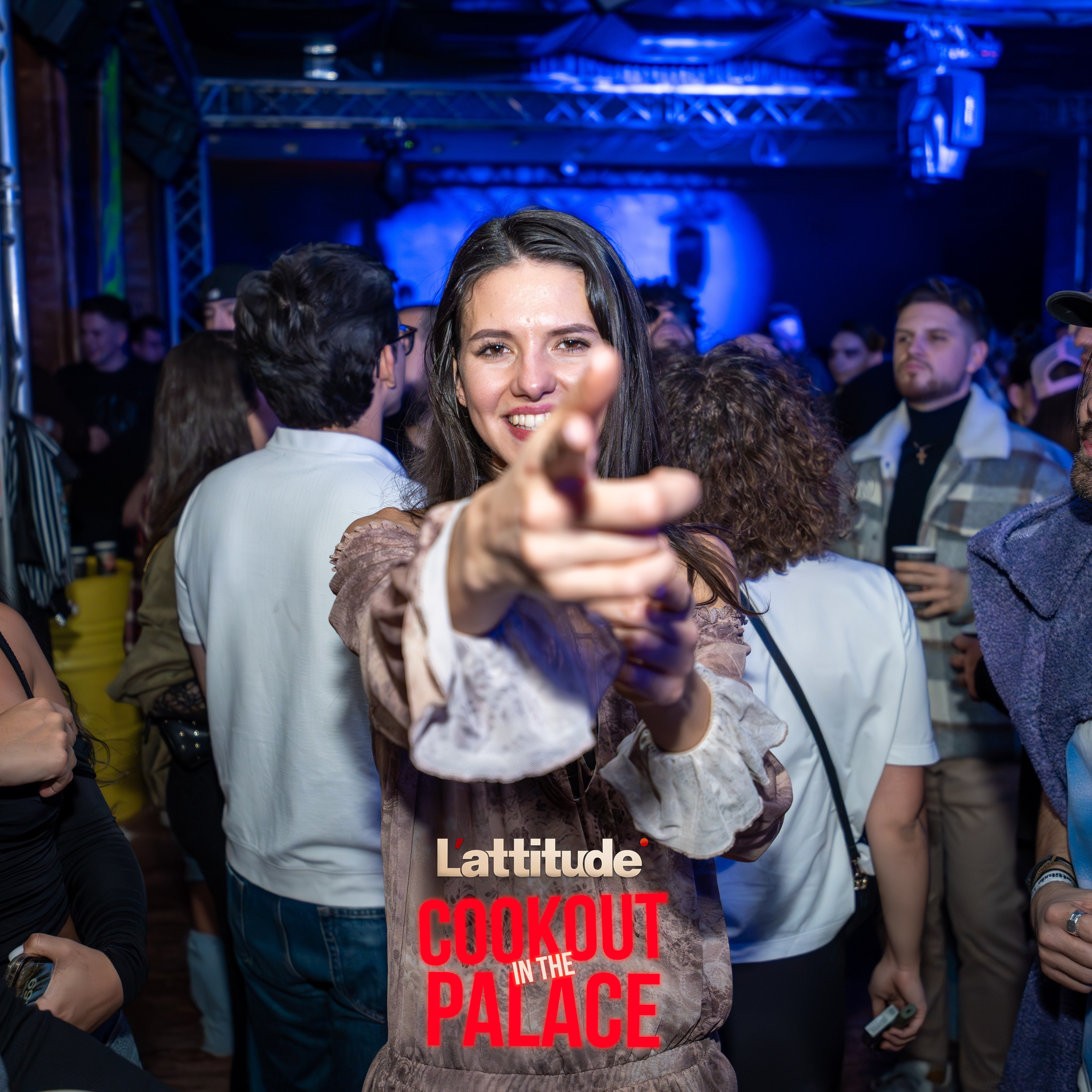 Young woman smiling and pointing towards the camera at a crowded indoor event with blue lighting.