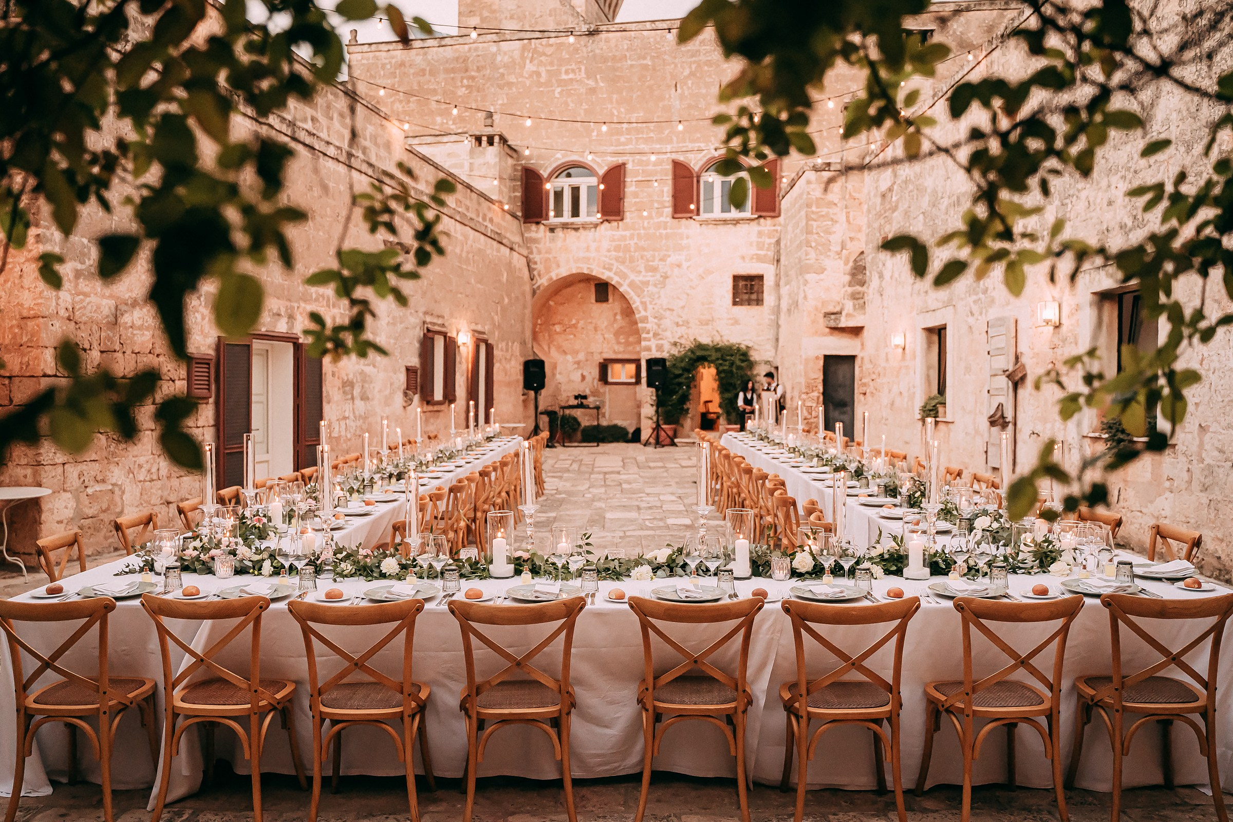 A beautifully arranged reception table in a historic courtyard, adorned with elegant white linens, greenery garlands, and candlelit ambiance.