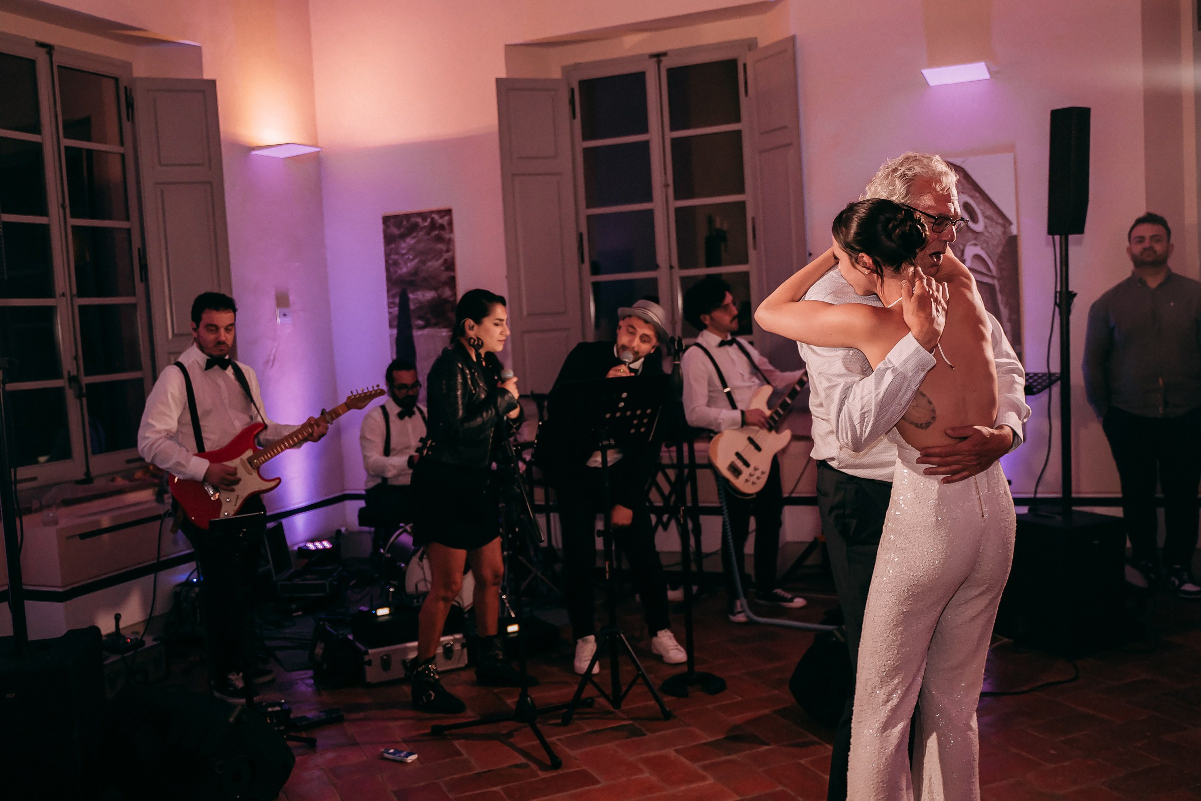 A bride and her father share an emotional embrace during a special dance, with a live band playing softly in the background of a warmly lit room.