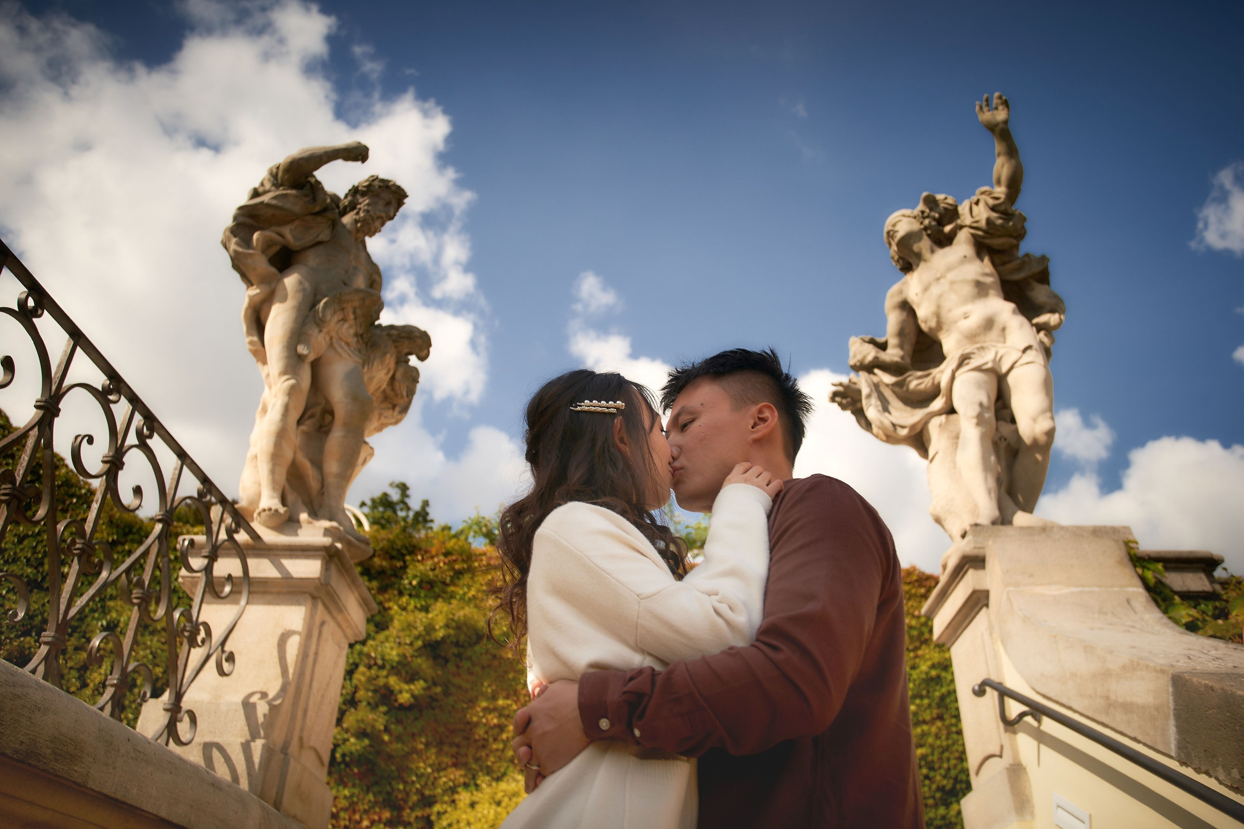 Eva and Conan kissing, framed by statues in Vrtba Garden against a blue sky.