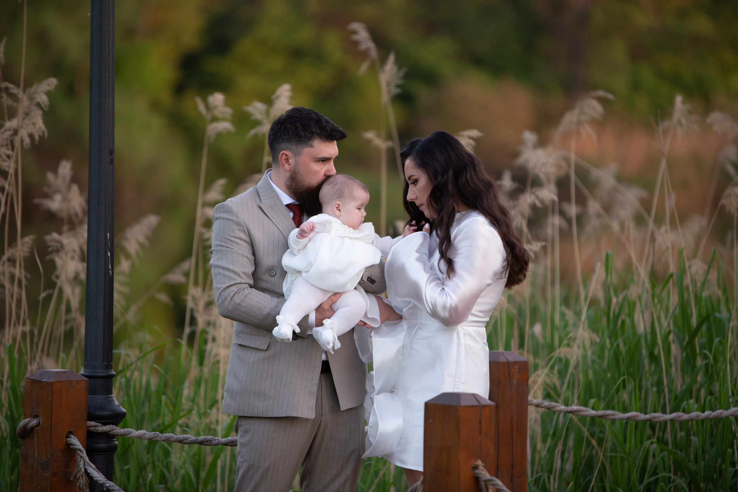 Man holding baby while woman looks at the baby outdoors against green foliage background.