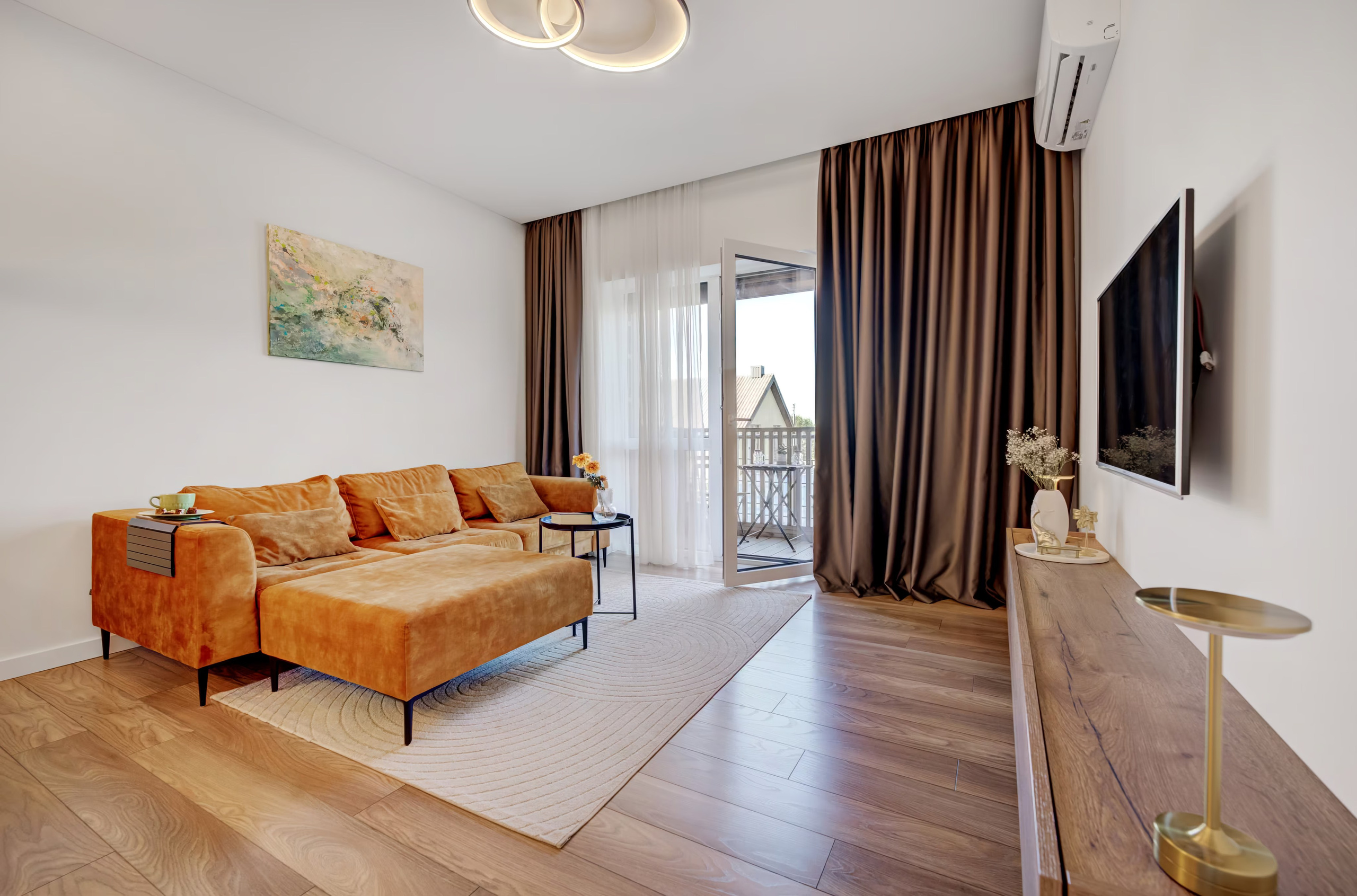 Jaukus gyvenamasis kambarys su aksomine oranž spalvos sofa, kilimu ir meno kūriniu ant sienos.