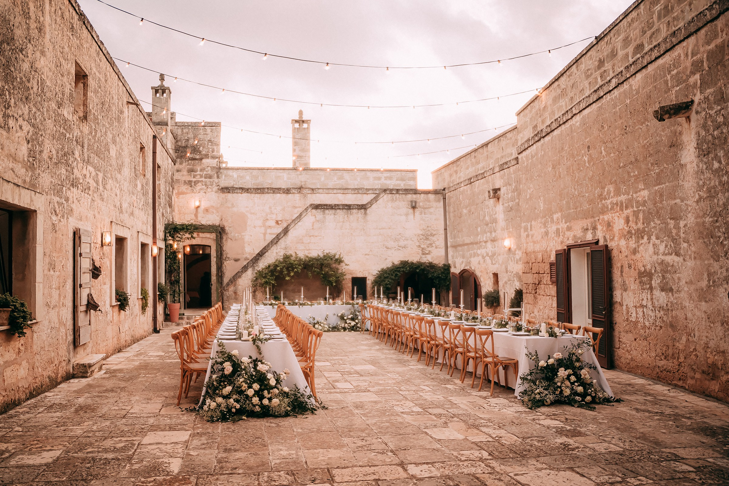 The courtyard of Masseria Salamina glows under string lights as dusk falls, with two banquet tables beautifully prepared for an evening celebration.
