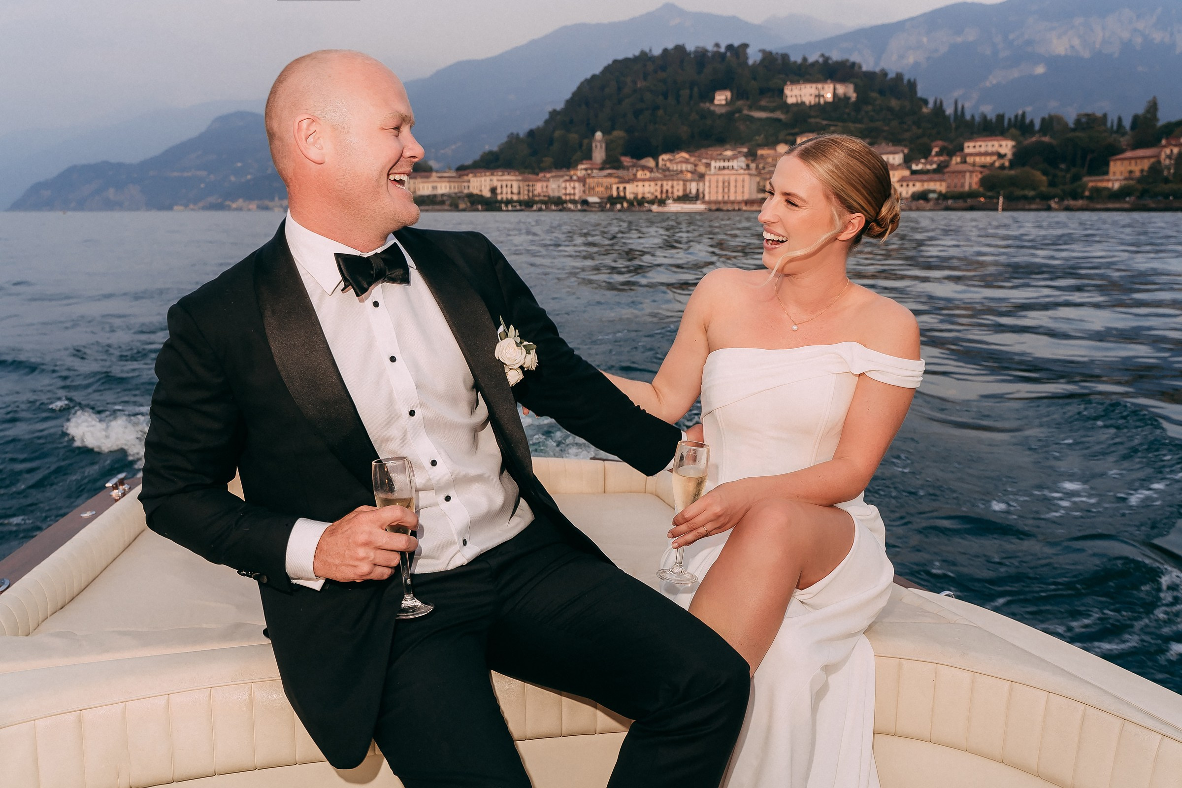 The couple toasts to their union with champagne on a boat, sharing a joyful and intimate moment surrounded by tranquil waters.