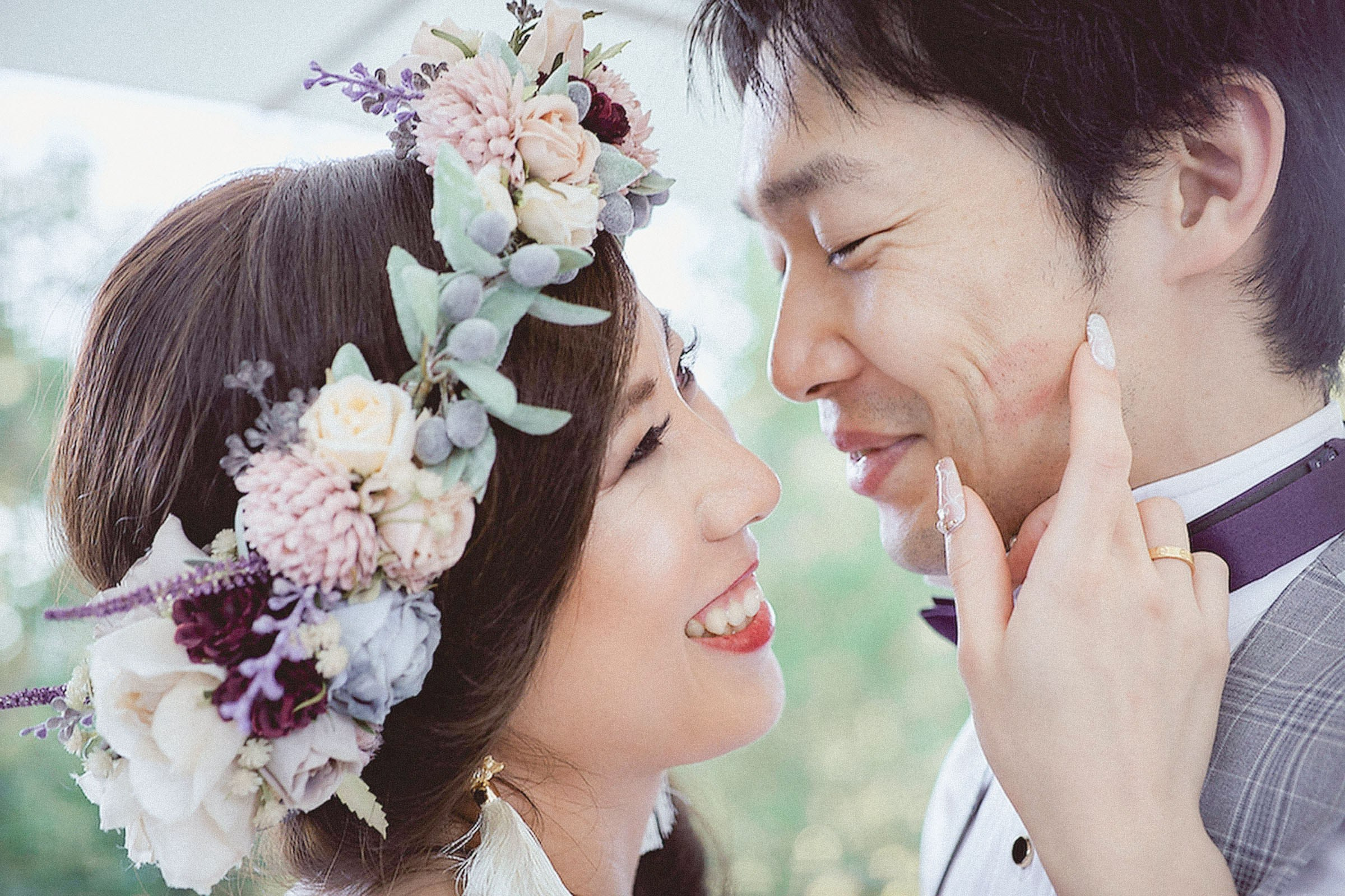 A smiling Japanese bride, wearing a floral headpiece lovingly strokes the face of her groom.