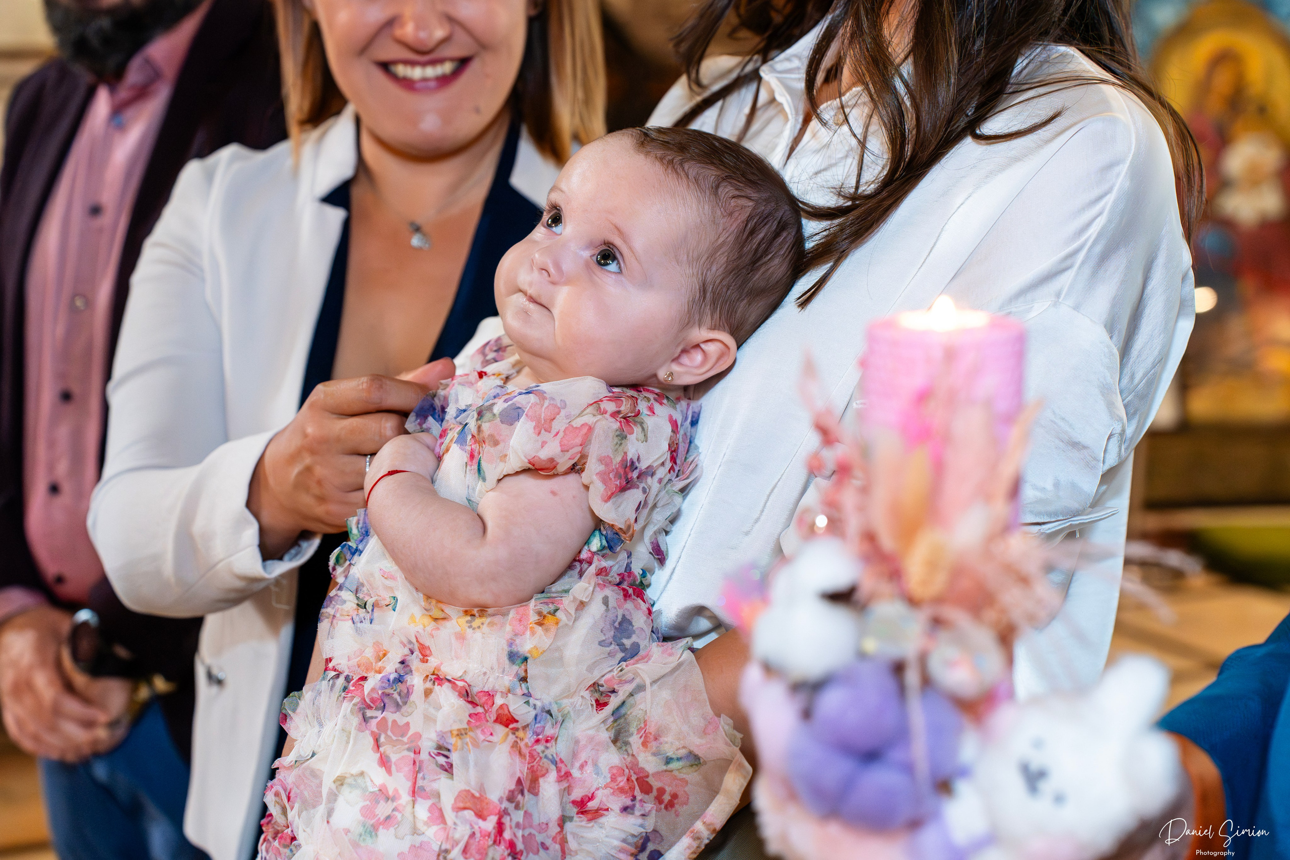 Baby in floral dress held by an adult woman during baptism ceremony with candle and decorations nearby.