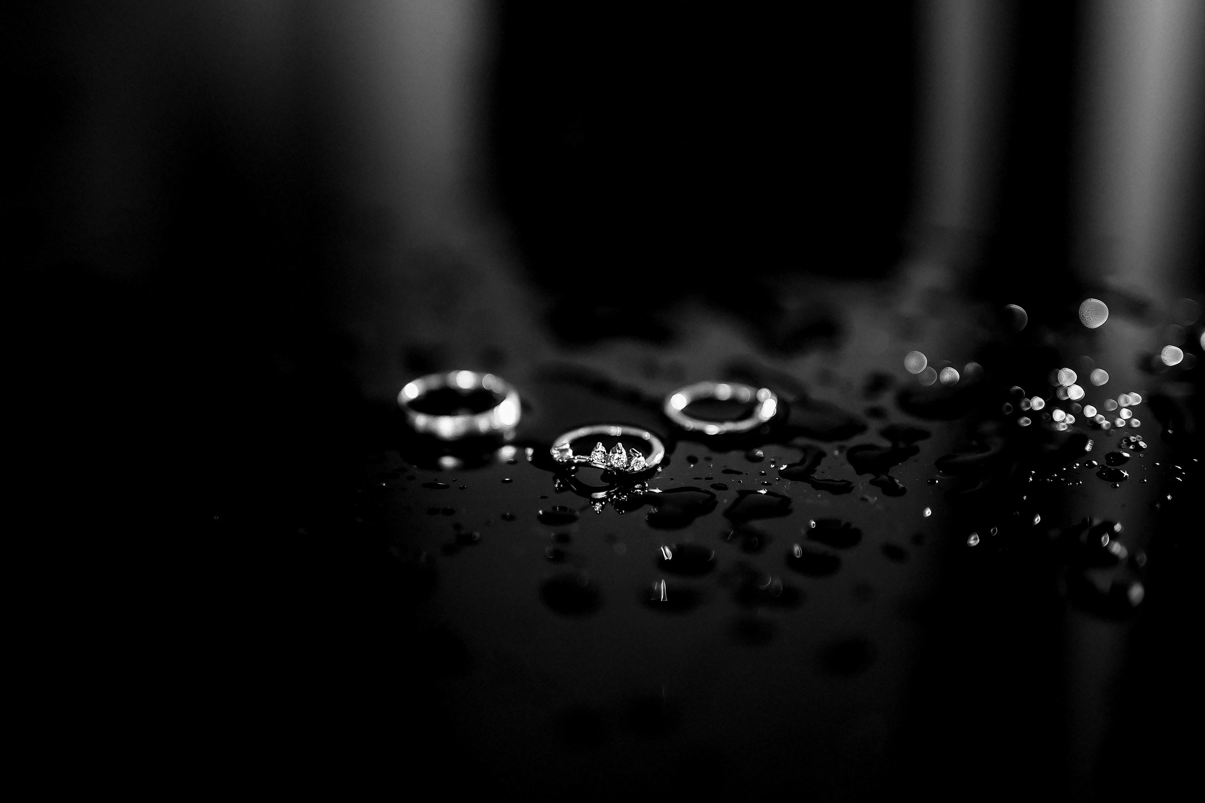 A close-up of three elegant rings on a black reflective surface, glistening with delicate water droplets.