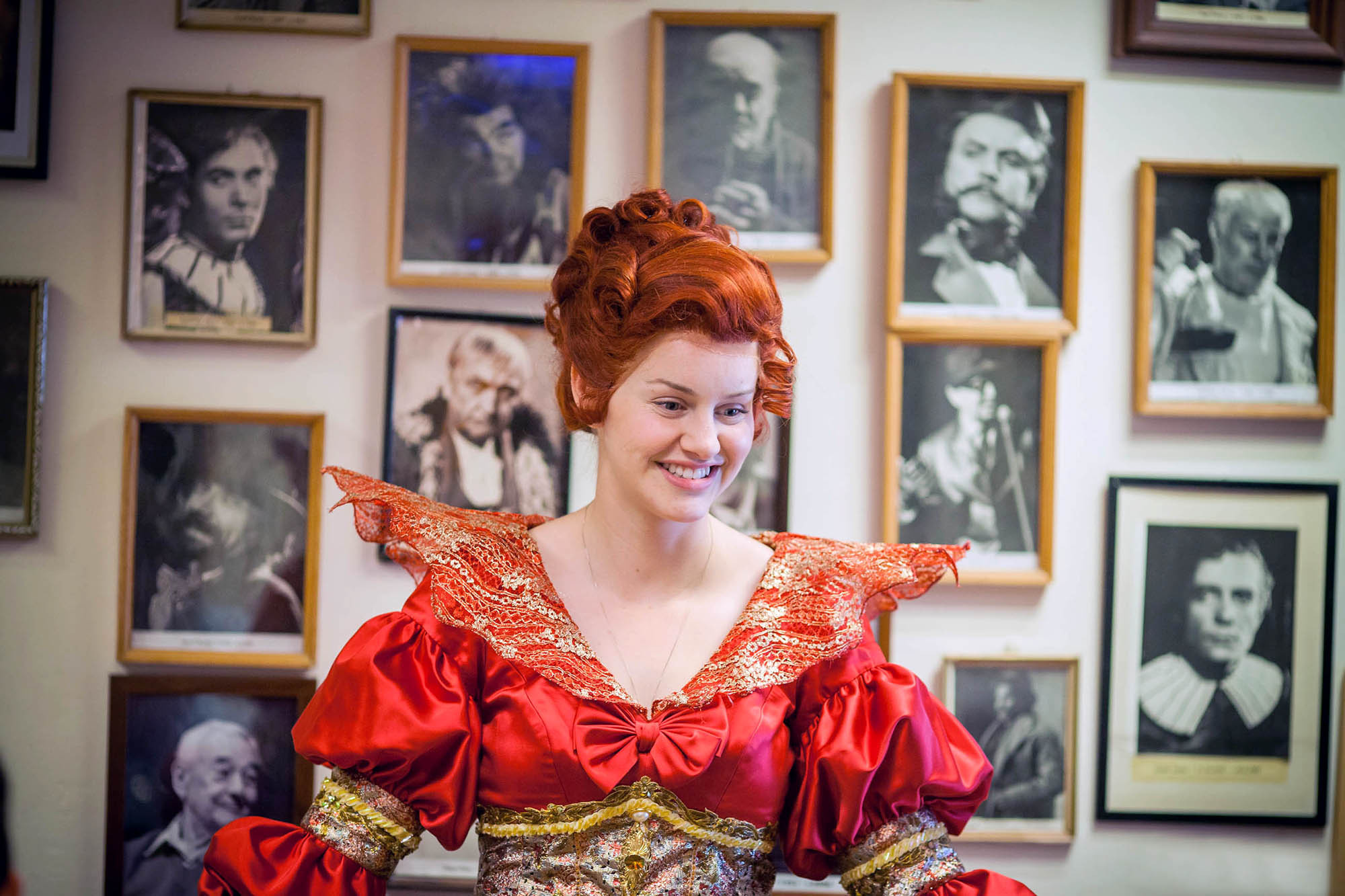 A behind the scenes photo of a smiling opera singer in medieval dress as she stands in front of a wall full of black and white portraits of stage performers at the Estates Theatre in Prague.