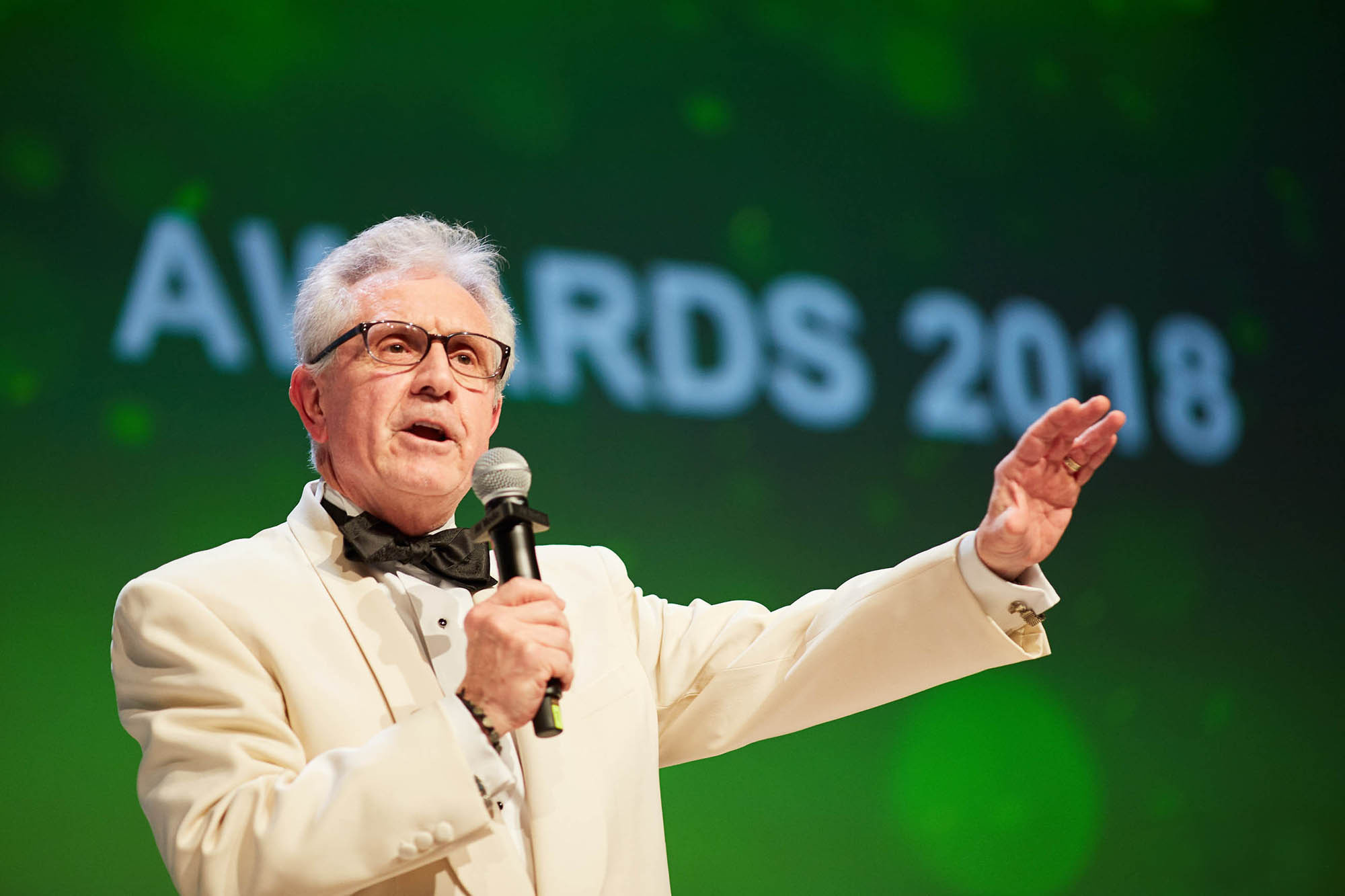 An older gentleman wearing a white suit and a black bow tie gestures on stage during an award ceremony.
