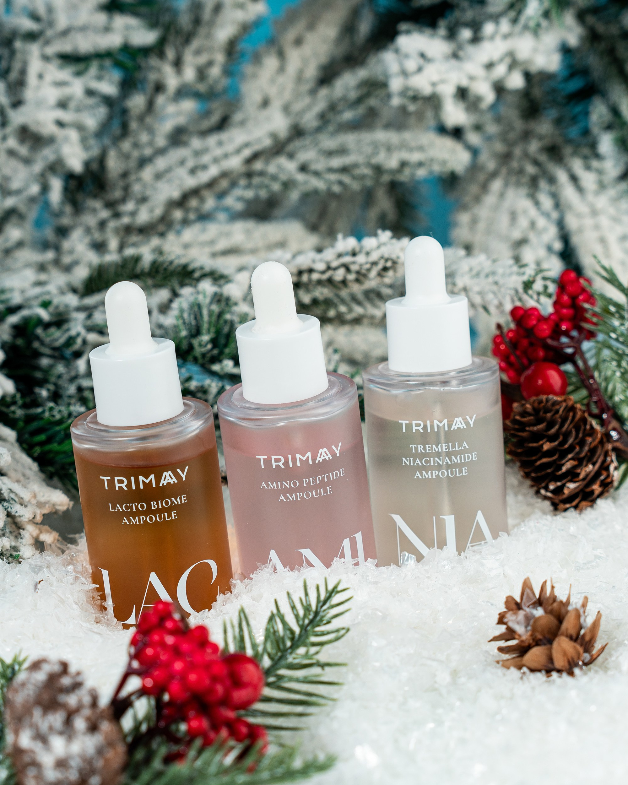 Qar, qozalar və qırmızı giləmeyvə bəzəkləri ilə əhatə olunmuş, soyuq, mavi fonda Trimay markalı üç damcı başlıqlı ampula (Lacto Biome, Amino Peptide və Tremella Niacinamide)