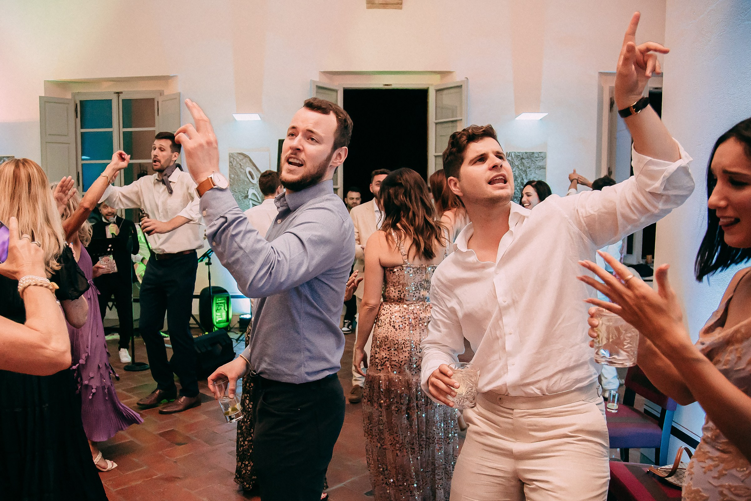 Two men enthusiastically pointing and singing along to the music on the dance floor, while others join in the festive atmosphere.