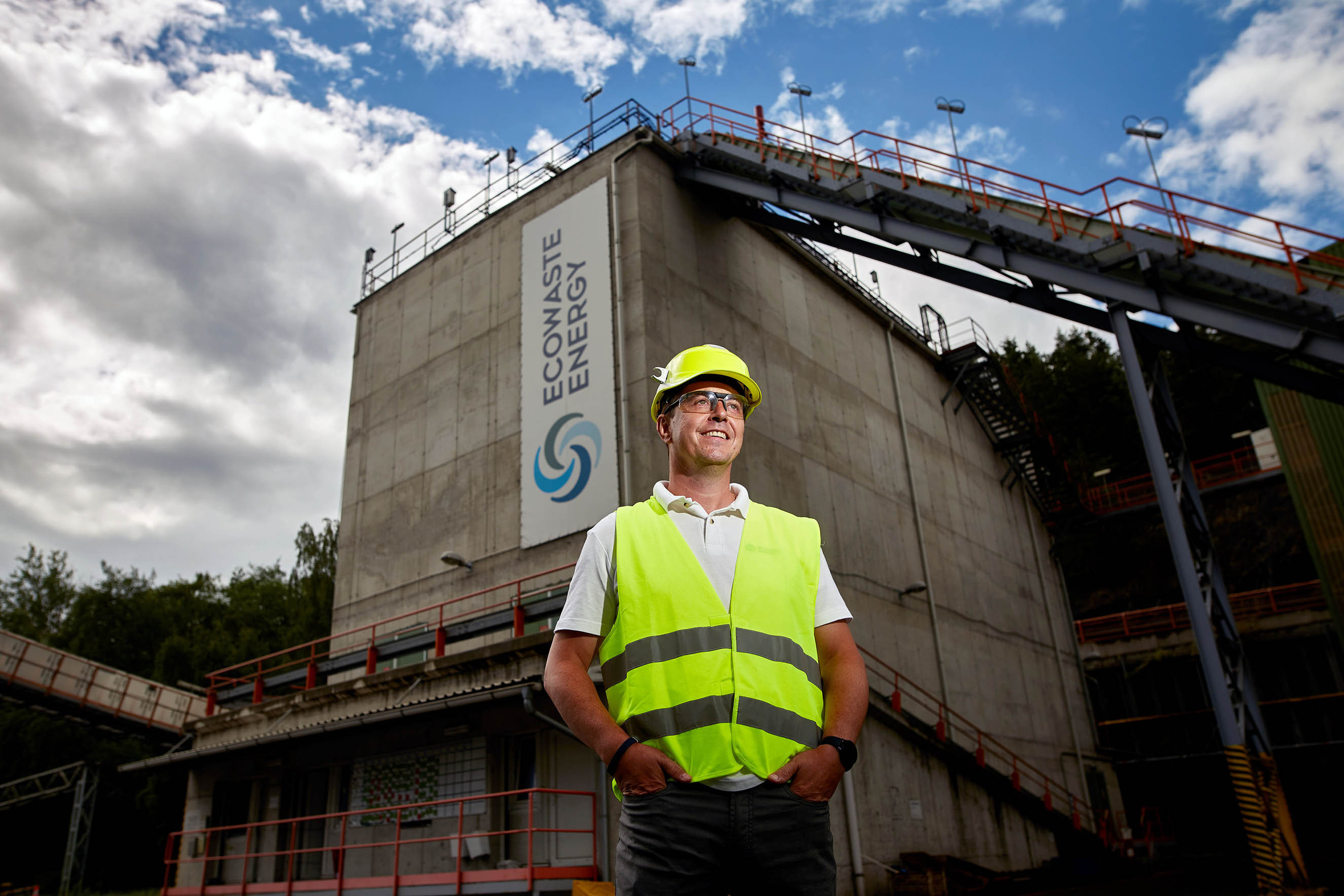 A man wearing a yellow reflective safety vest and hard hat looks away from the camera as the Ecowaste Energy plant is seen directly behind him in Prachovice, Czech Republic.