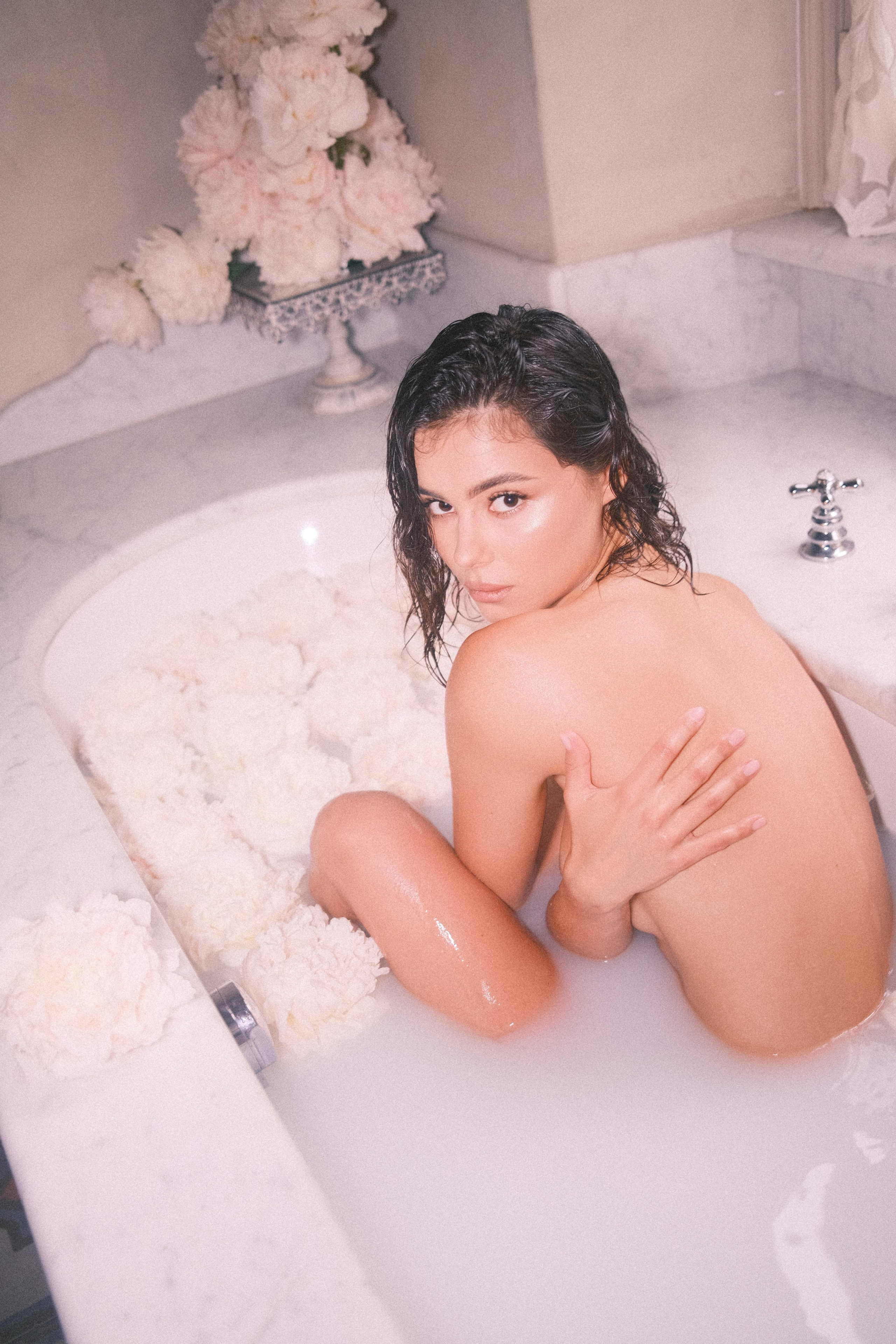 A serene shot of a woman in a white marble bathtub filled with milky water and surrounded by pale pink flowers, her gaze captivating as she looks over her shoulder.