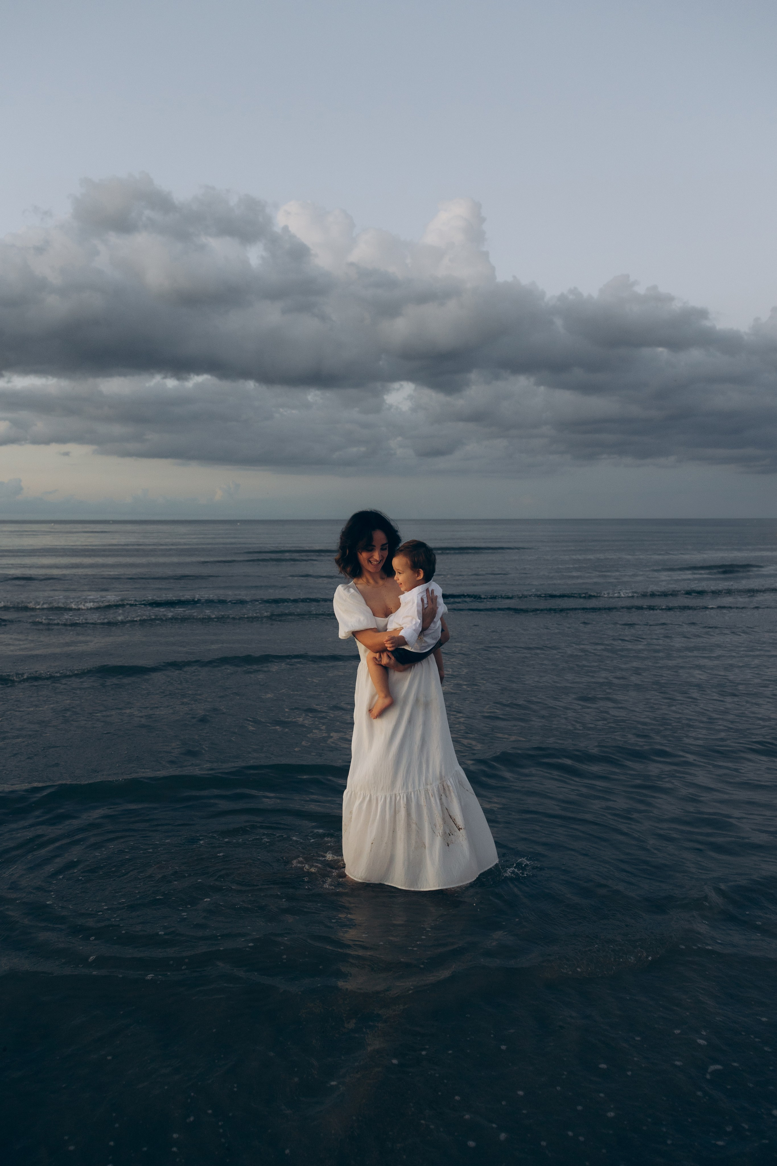 Madre e bambino nell’acqua a Rimini con cielo nuvoloso sullo sfondo. Fotografia di famiglia artistica che racconta emozione e connessione.