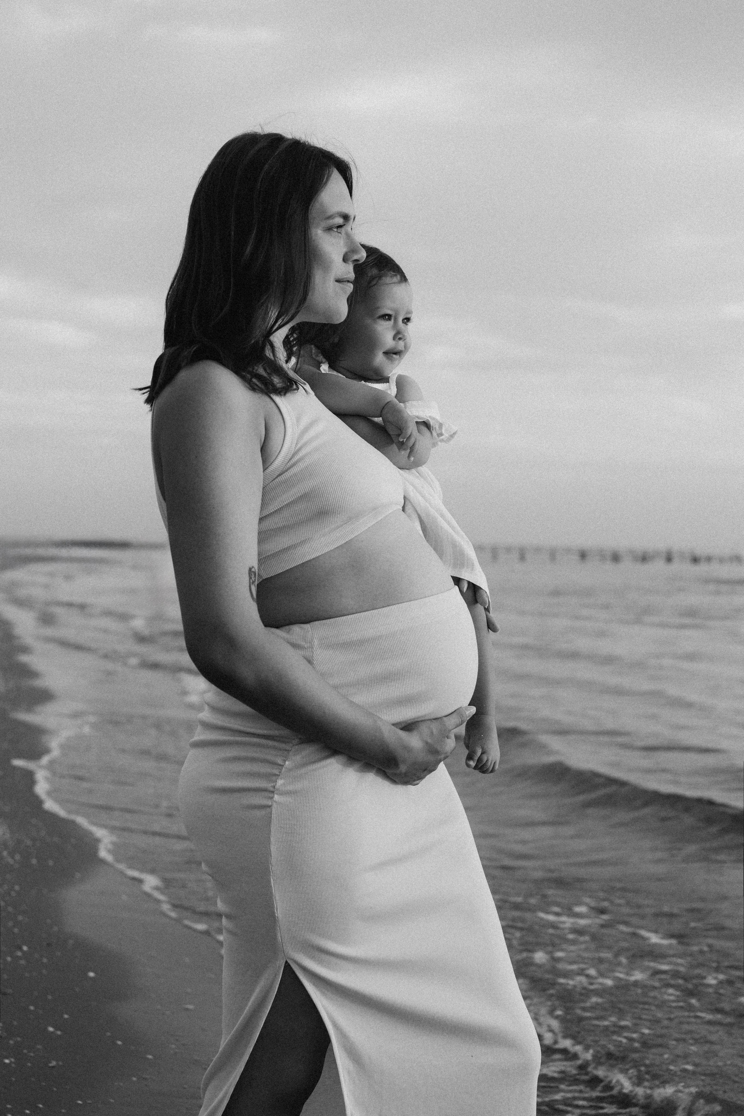Ritratto di famiglia in bianco e nero sulla spiaggia di Bellaria: una donna incinta cammina lungo la riva del mare Adriatico tenendo in braccio una bambina, le onde leggere e l’orizzonte aperto raccontano un momento intimo di maternità, attesa e legame familiare.