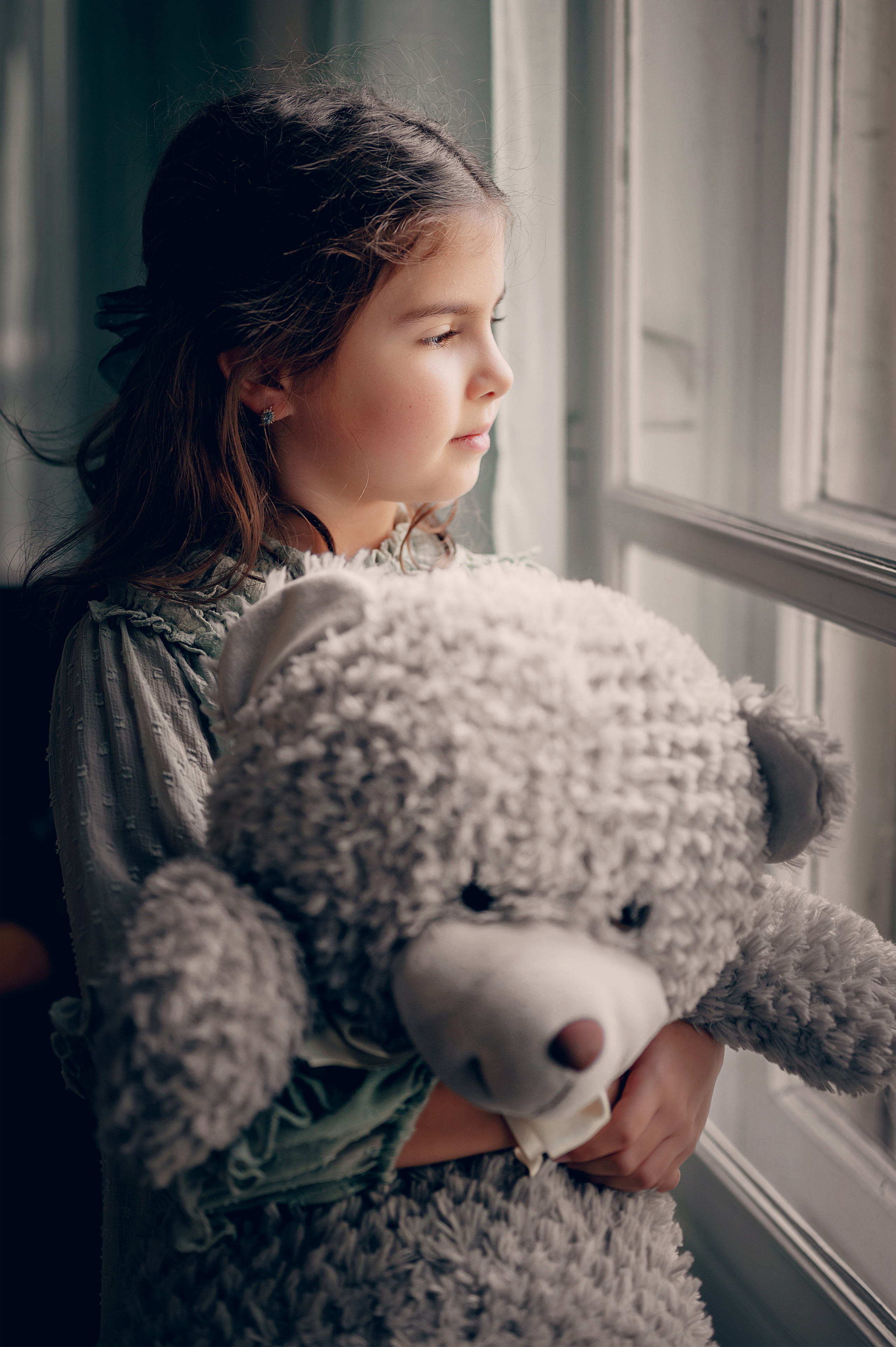 Une fille avec un jouet en peluche regarde par la fenêtre, plongée dans ses pensées.