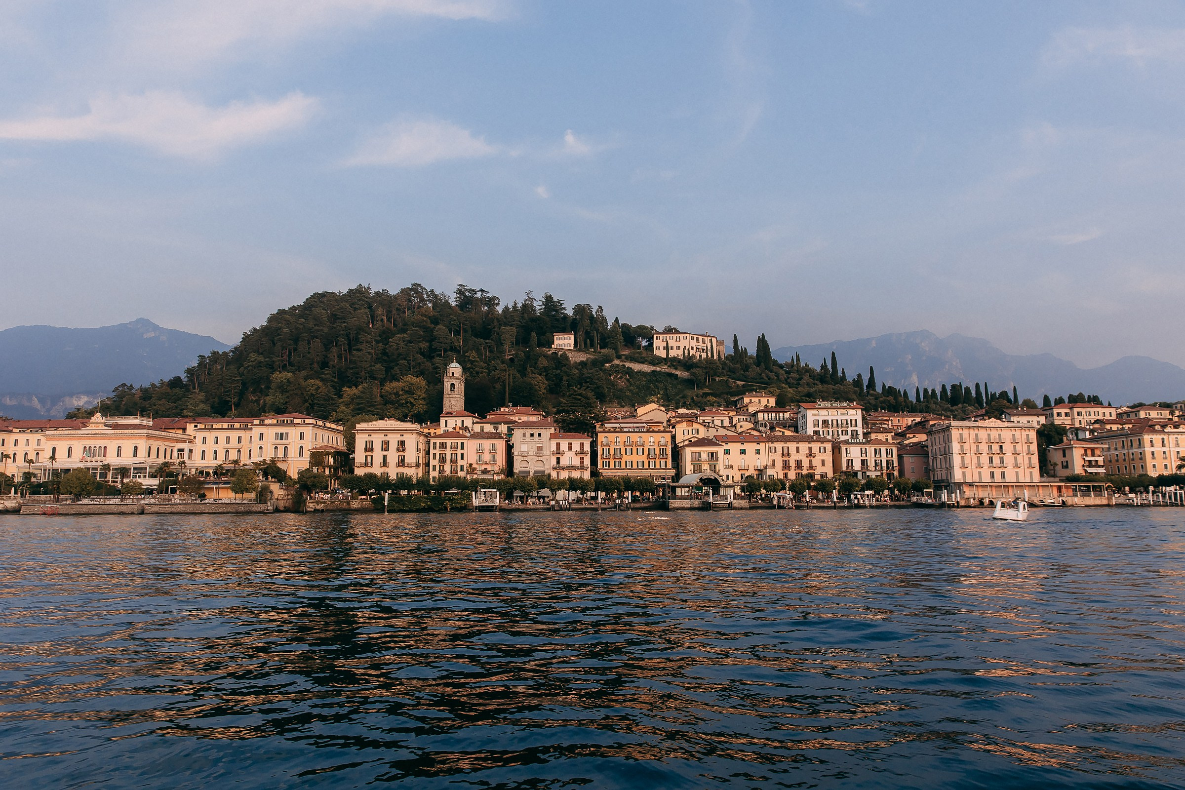 A stunning view of a picturesque lakeside town with historic buildings nestled on a hill, surrounded by lush greenery and mountains in the background, reflected in the calm waters.