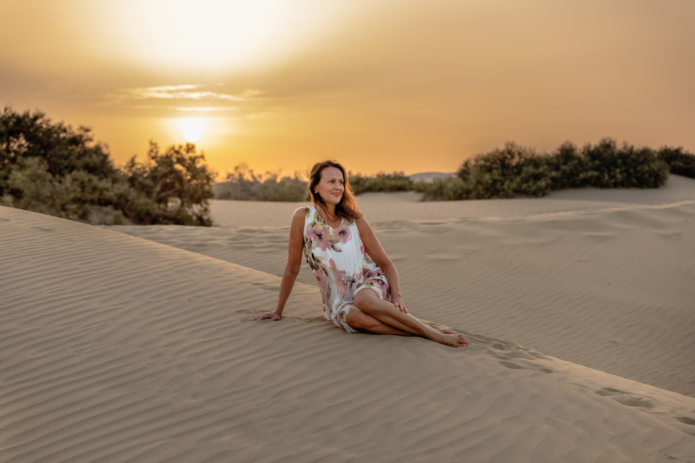 A woman sitting on a sand dune at sunset.
