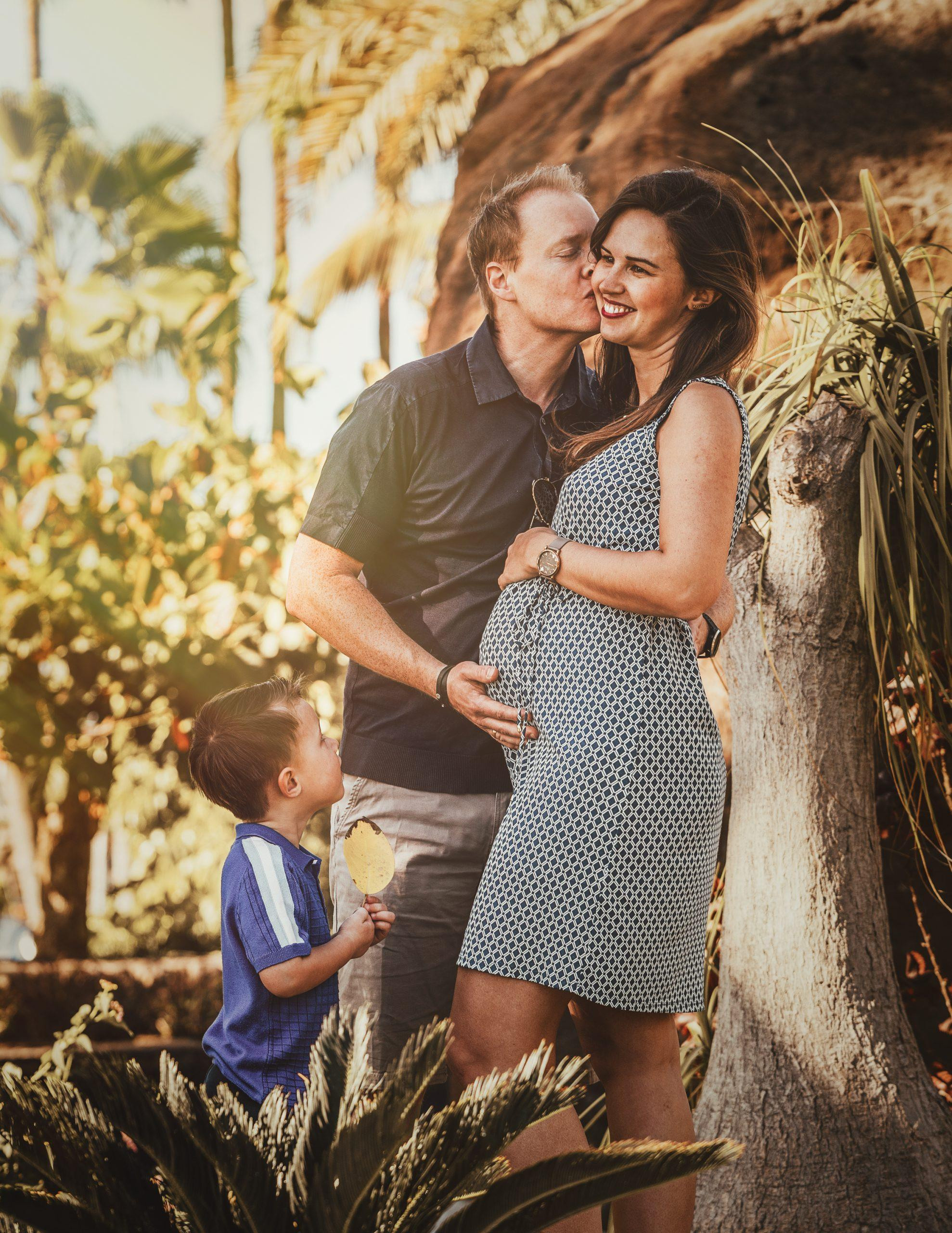 A pregnant couple kissing in front of a palm tree