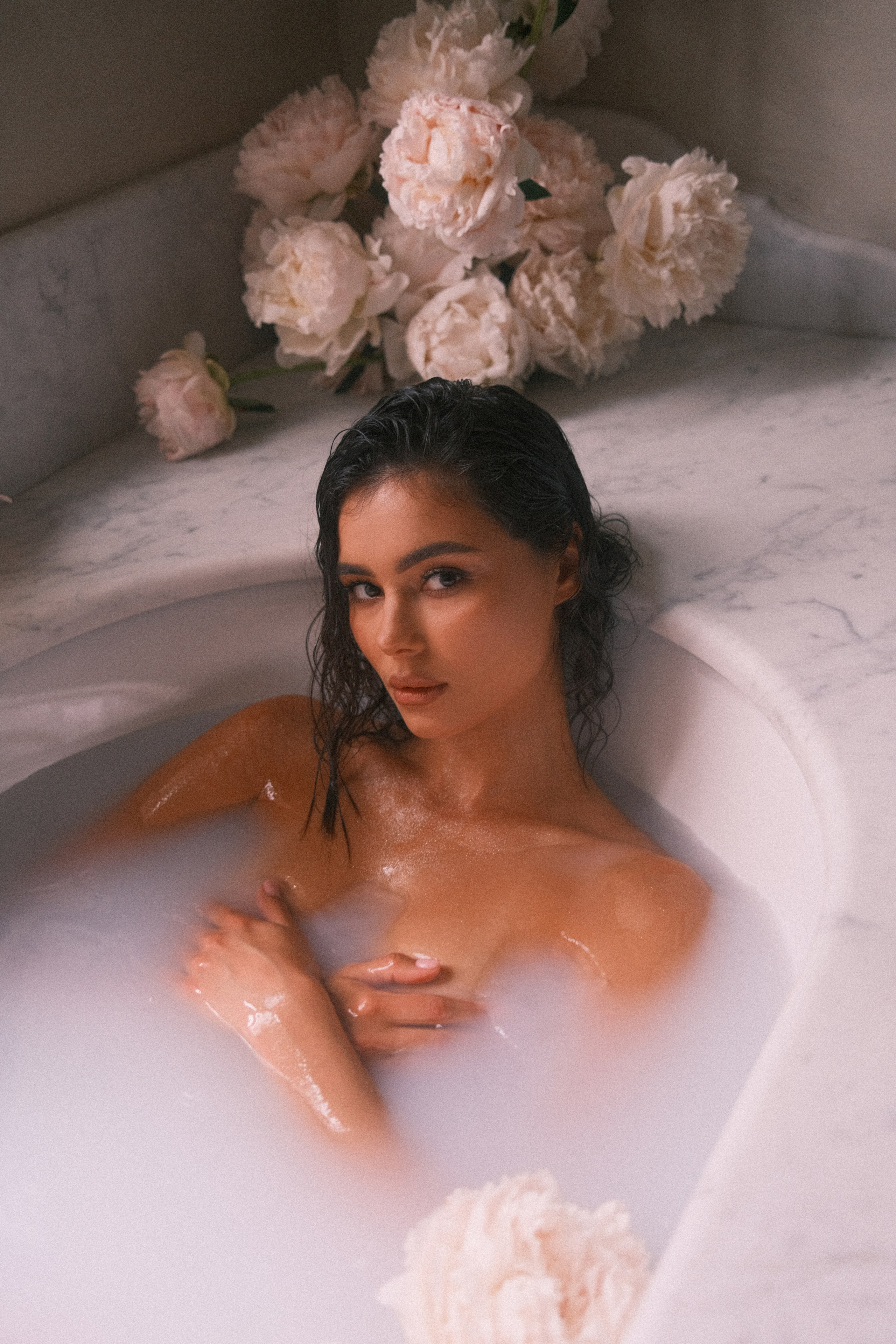 Close-up of a bride partially submerged in a milky bath, surrounded by soft pink flowers, gazing directly at the camera.