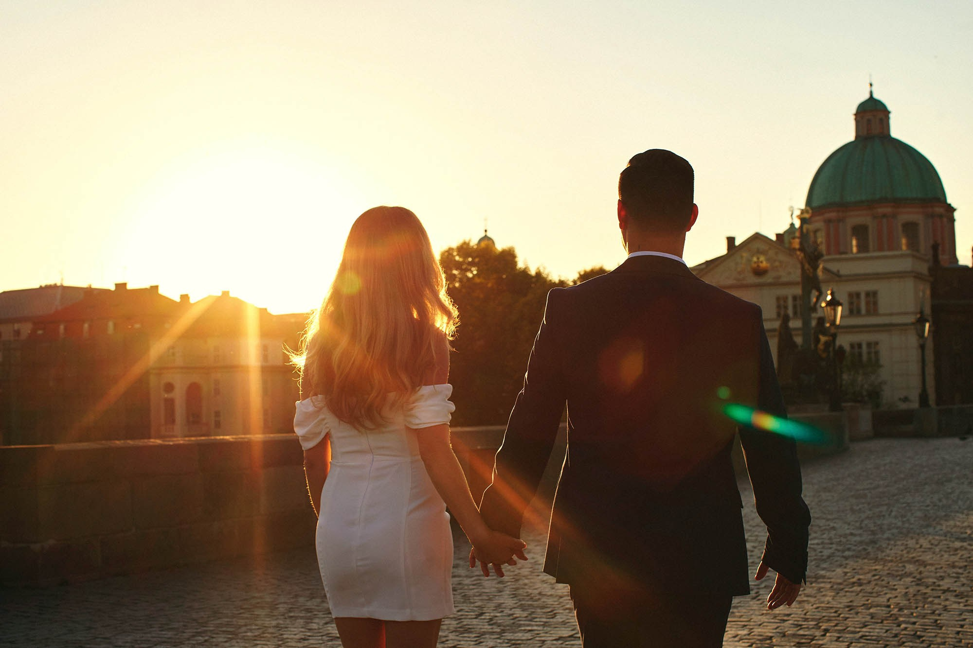 A couple walking across Charles Bridge with a dramatic sunrise flare over the skyline