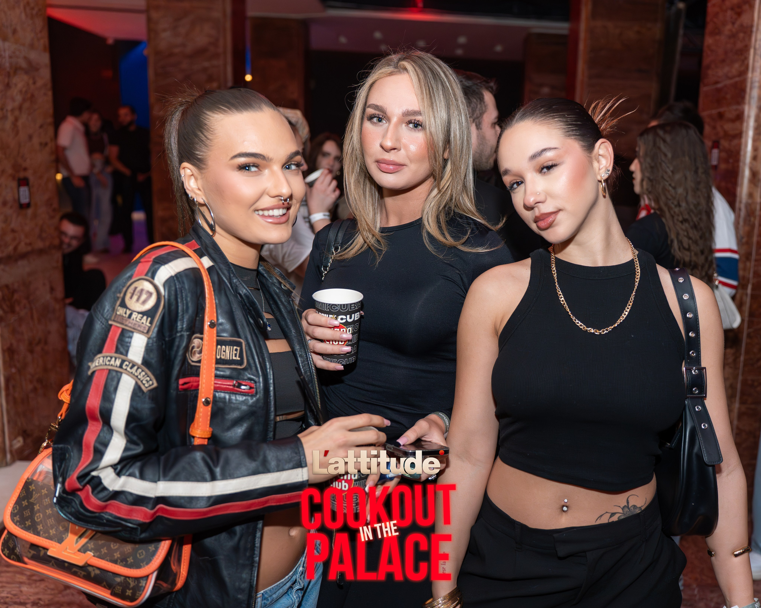 Three women posing at an indoor social event with people in the background.