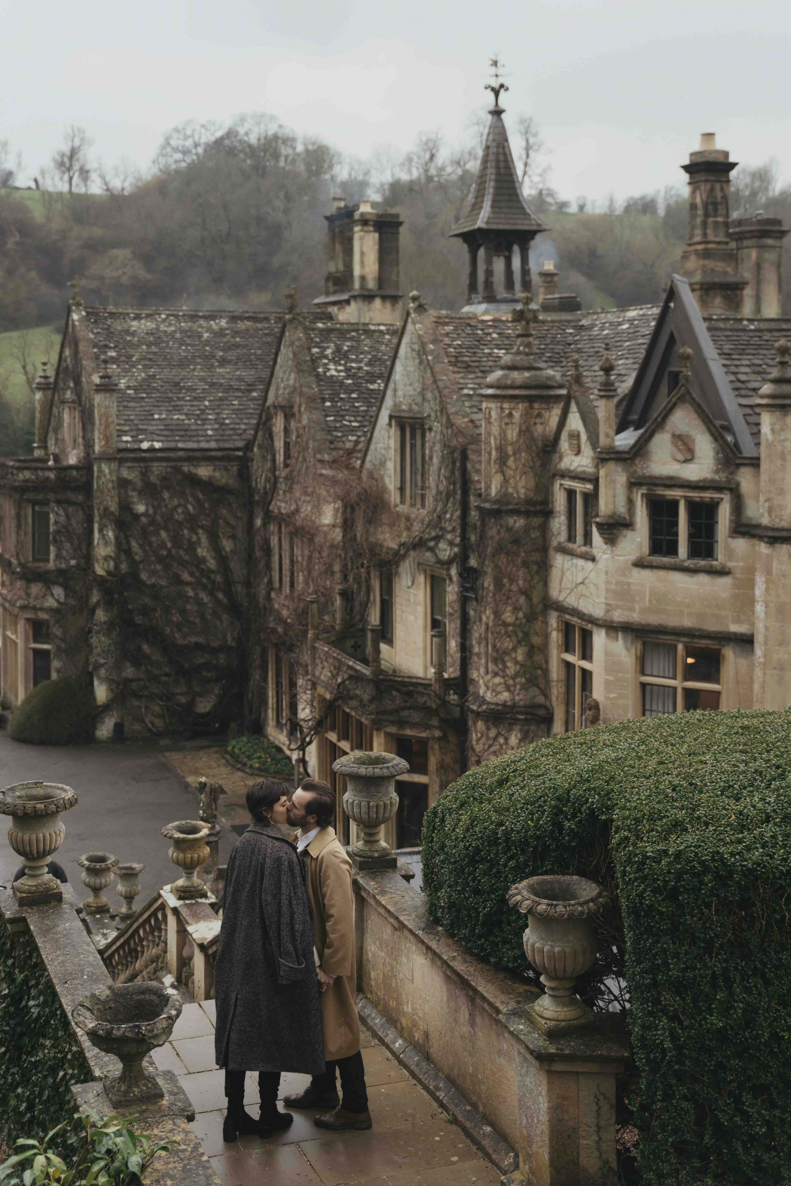 couple walking near historic manor house in Cotswolds countryside romantic engagement session