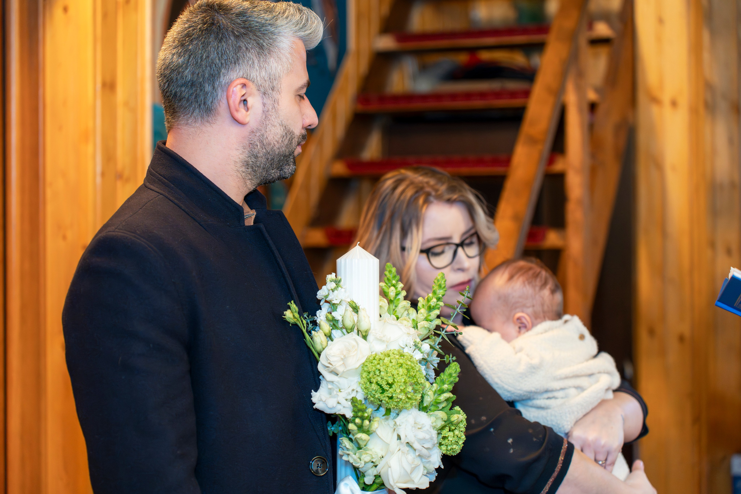 Man holding candle with flowers, woman holding baby indoors near wooden stairs.