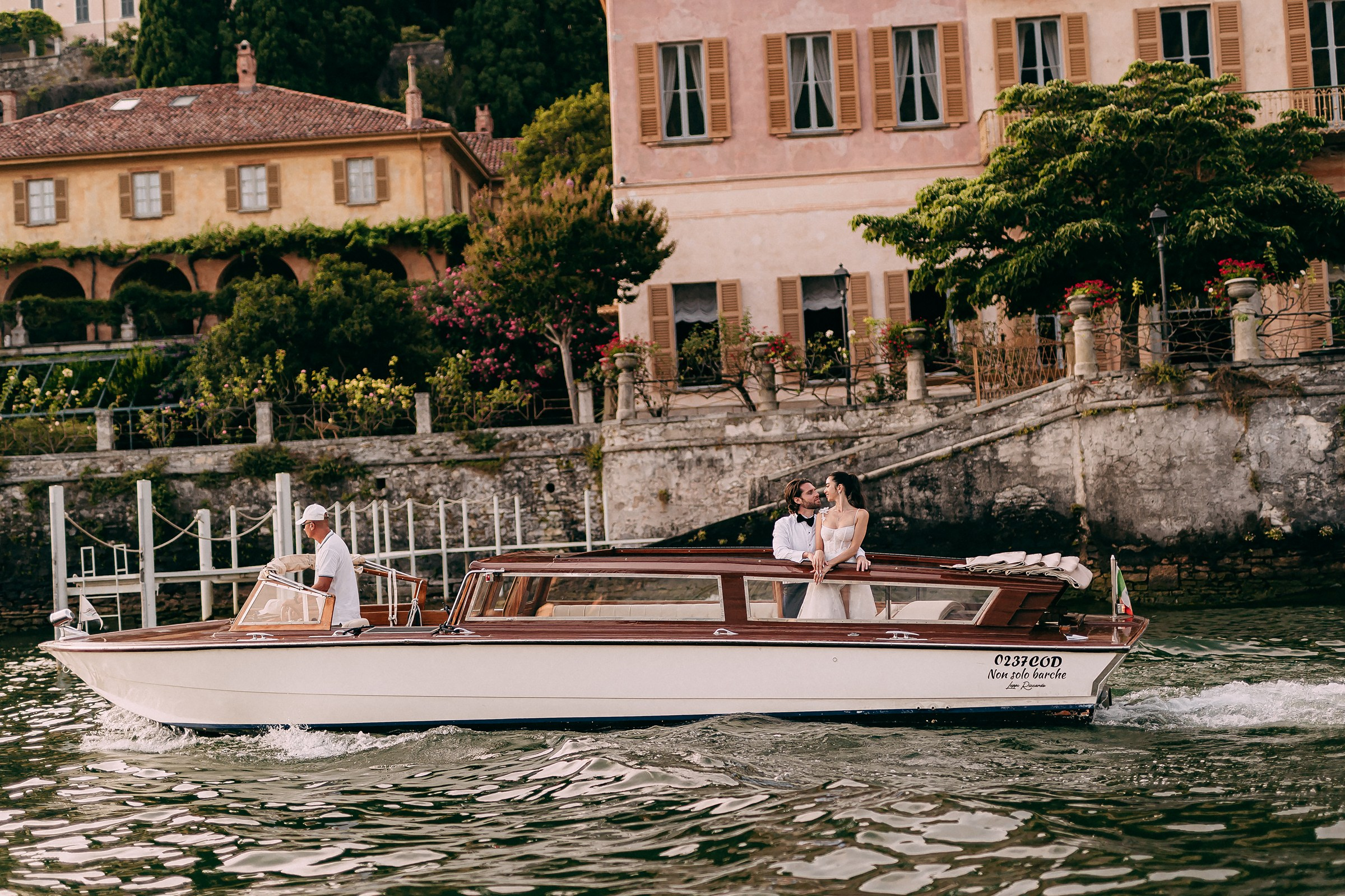 Boat glides past picturesque lakeside villas, with bride and groom enjoying the view.