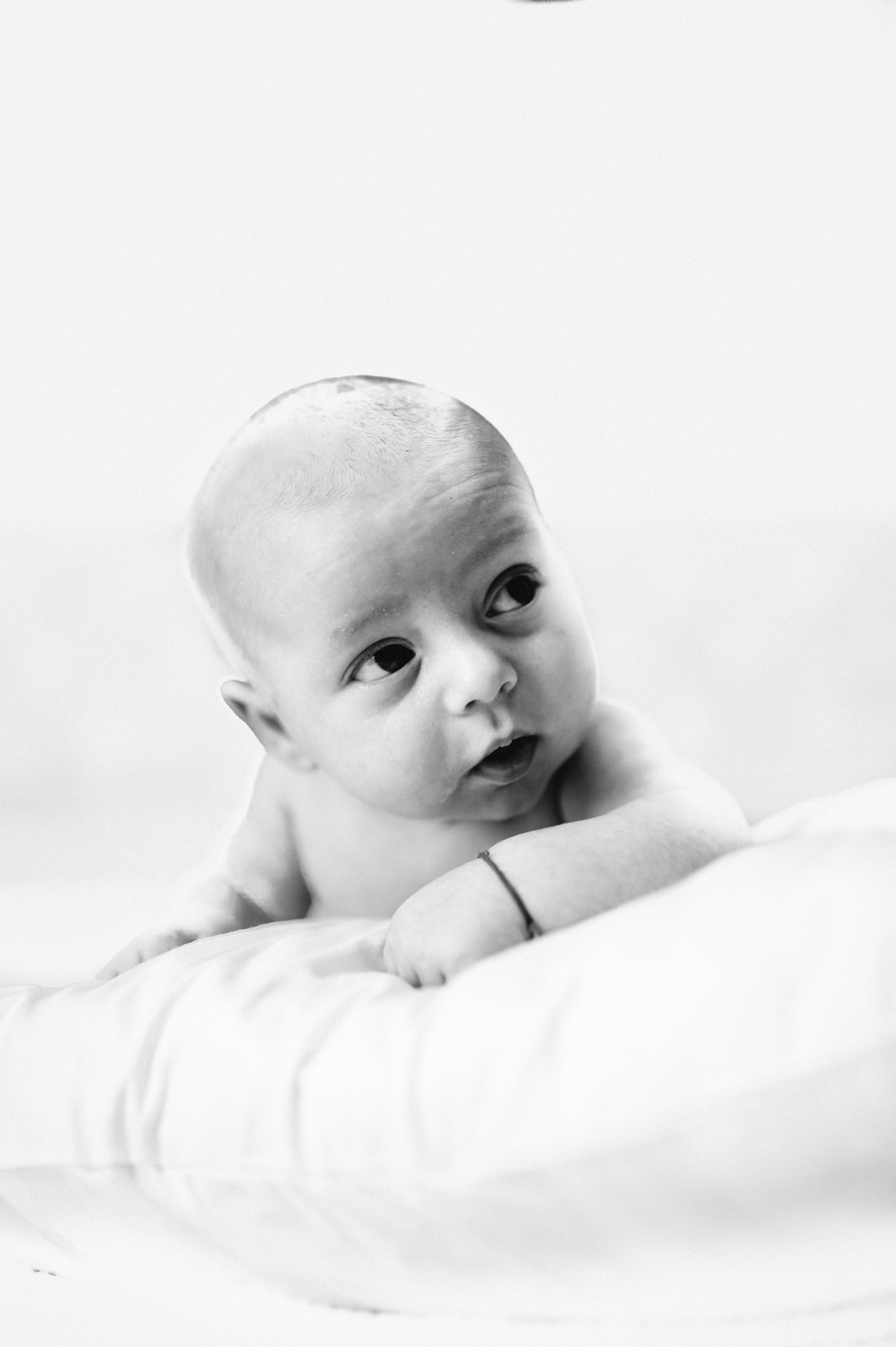 Portret alb-negru al unui bebeluș curios, sprijinit pe o pernă moale, privind atent în lateral.Chip inocent de bebeluș, surprins într-un moment de uimire și delicatețe.Bebeluș adorabil, cu ochii mari și curioși, odihnindu-se pe o pernă pufoasă.Moment tandru: bebeluș în alb-negru, descoperind lumea cu priviri pline de mirare.Portret artistic al unui nou-născut, evidențiind inocența și puritatea copilăriei.