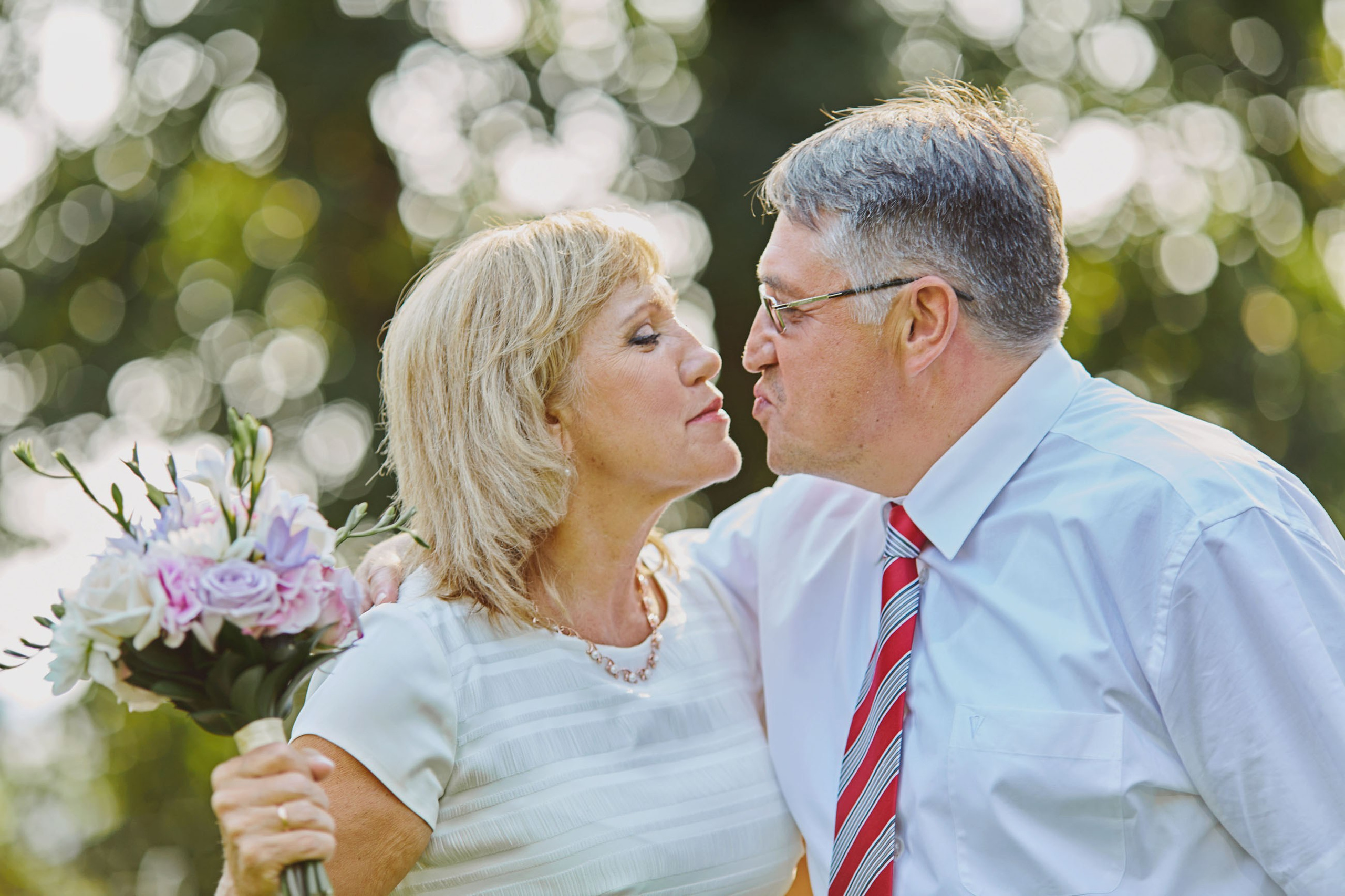 The groom's parents cuddle up and imitate a kiss as the mother holds a bouquet of flowers following their son's beautiful summer wedding.