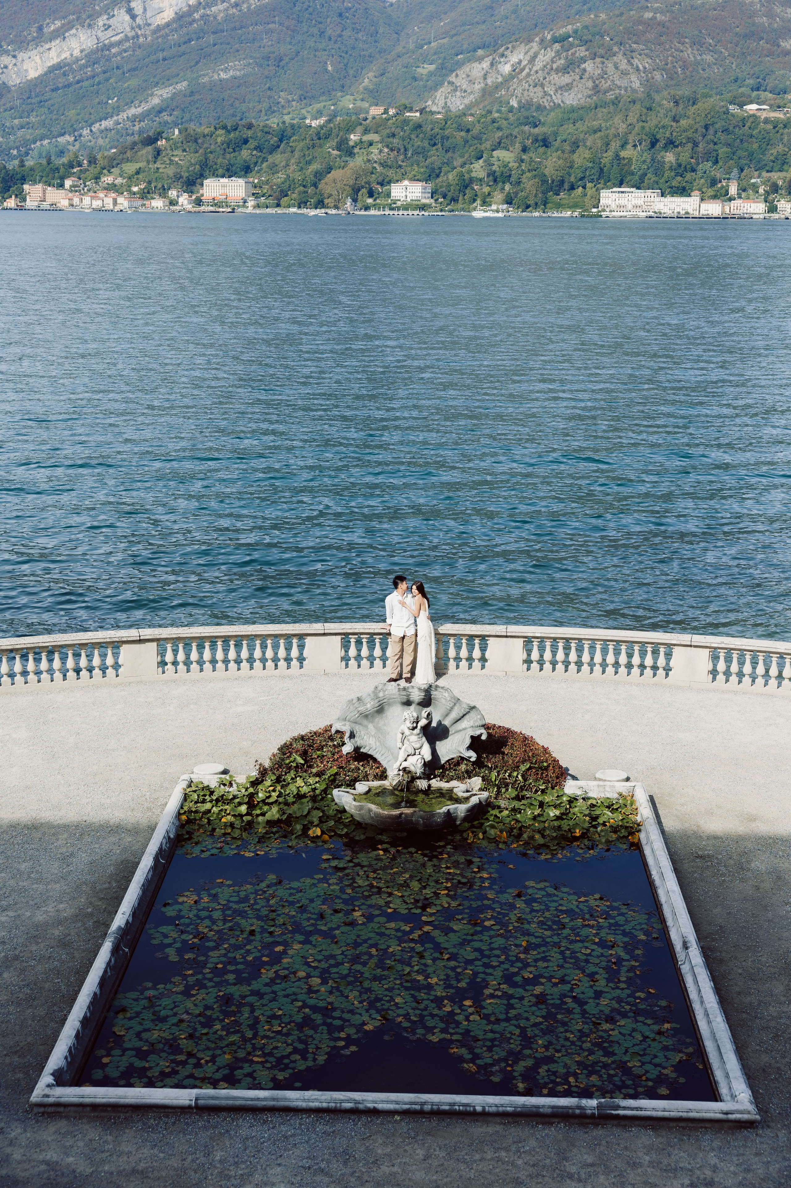 Couple embracing on the lakeside terrace of Villa Melzi, overlooking the blue waters of Lake Como.