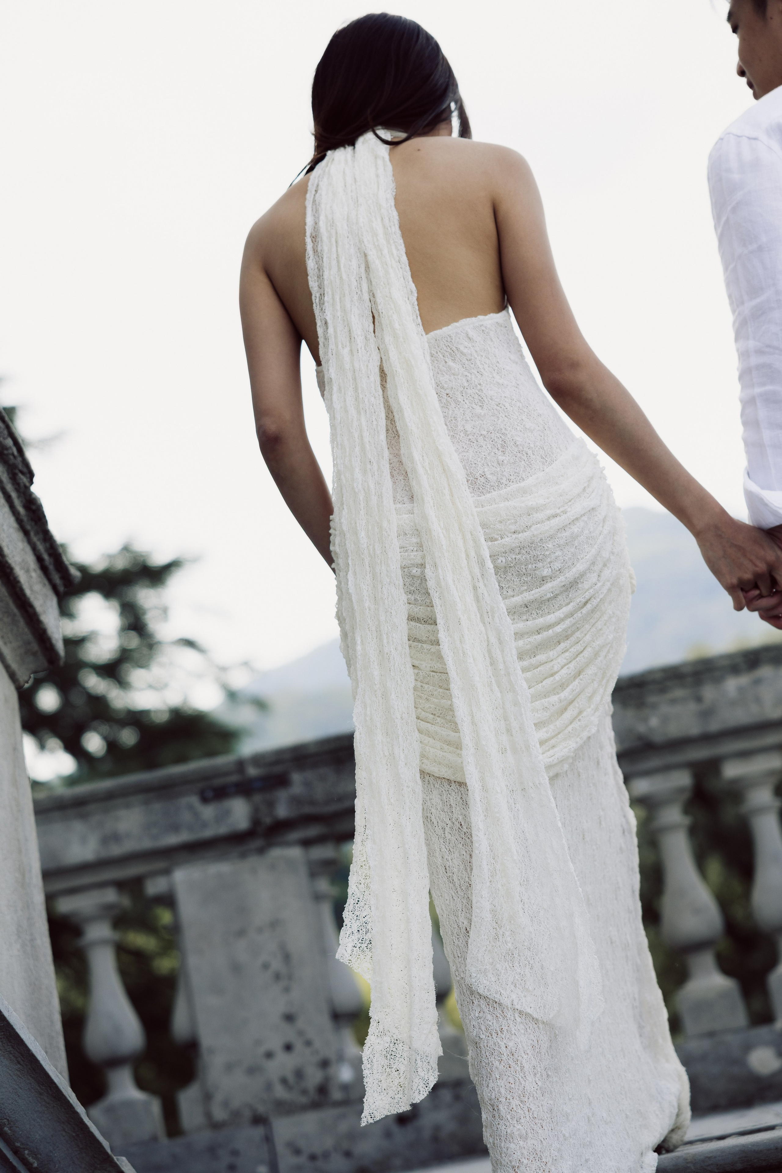 Woman in an elegant white dress holding her partner's hand beside a stone balustrade at Lake Como.