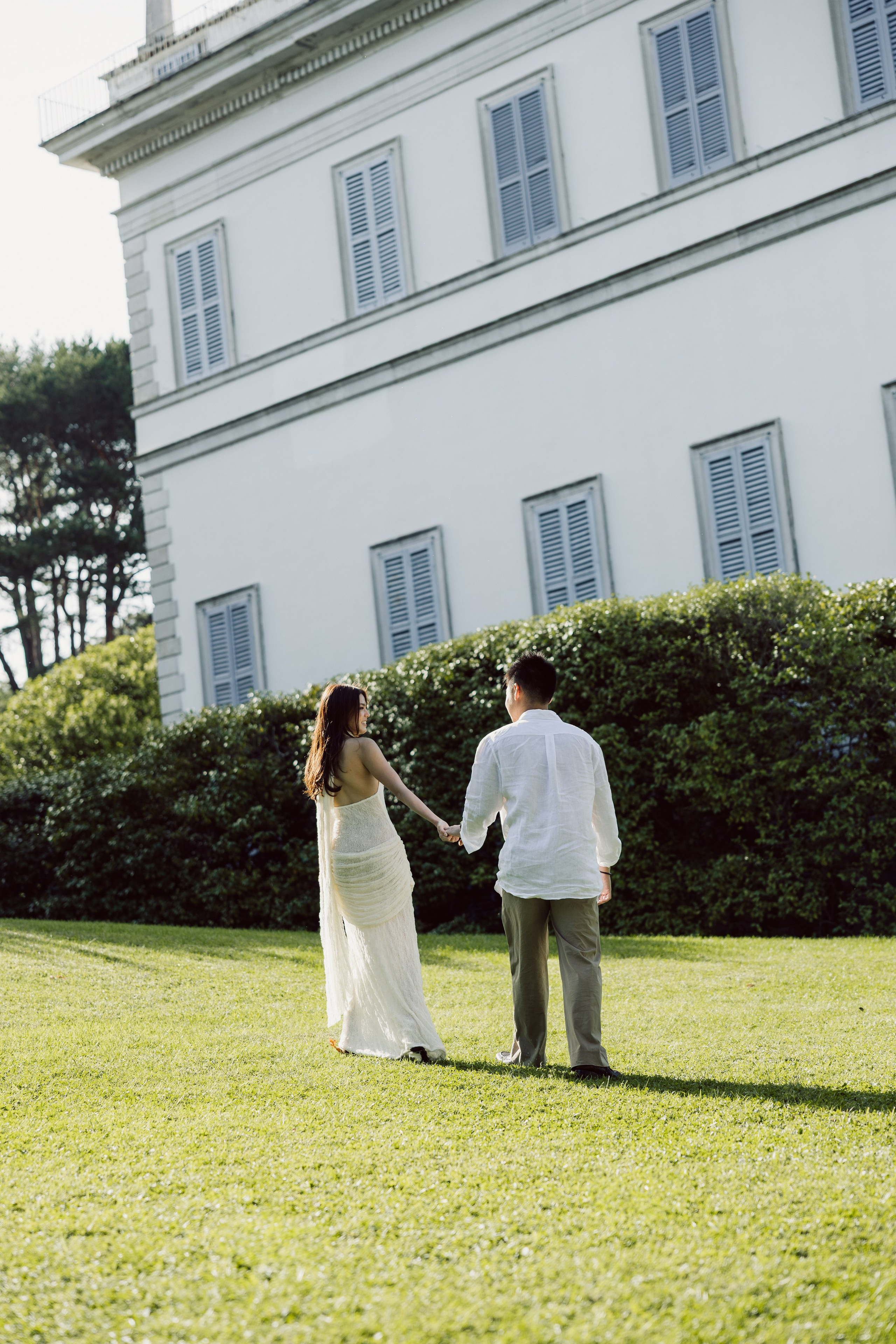 Couple holding hands on a sunlit lawn beside a historic villa in the gardens of Bellagio.