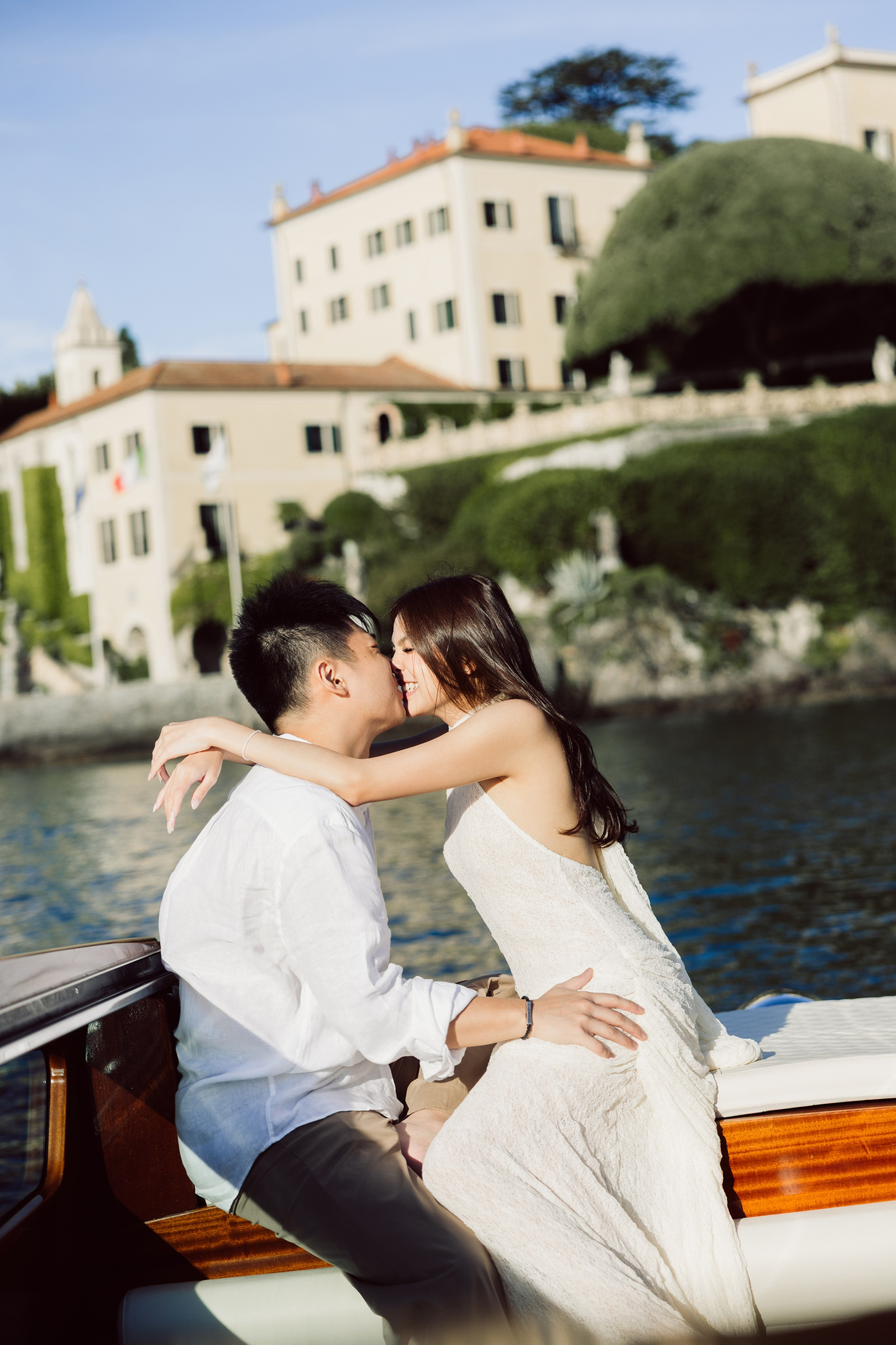Couple sharing a romantic kiss on a wooden boat with a lakeside Villa del Balbianello behind them