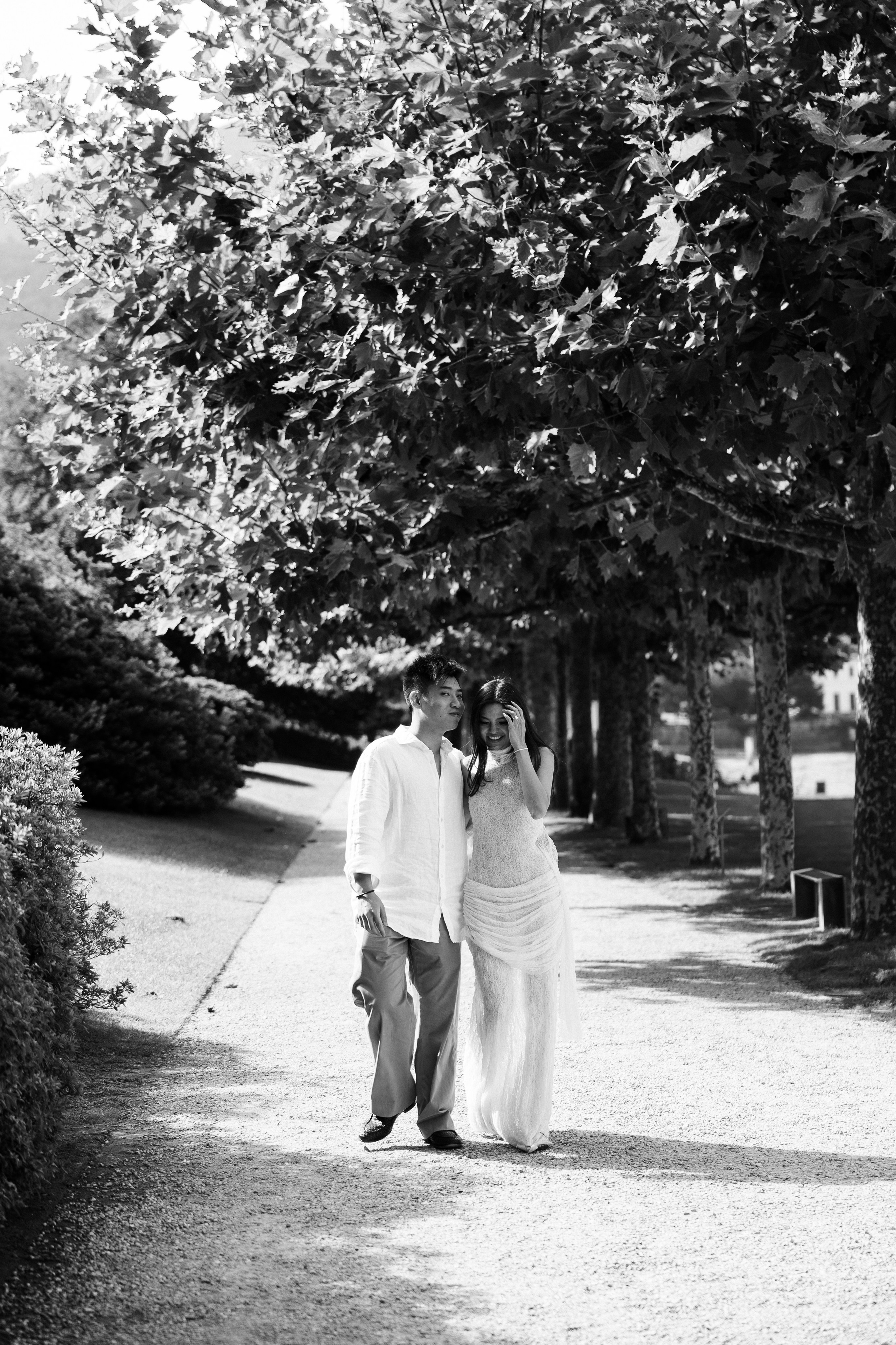 Black and white photograph of a couple walking together beneath the trees in the gardens of Bellagio.
