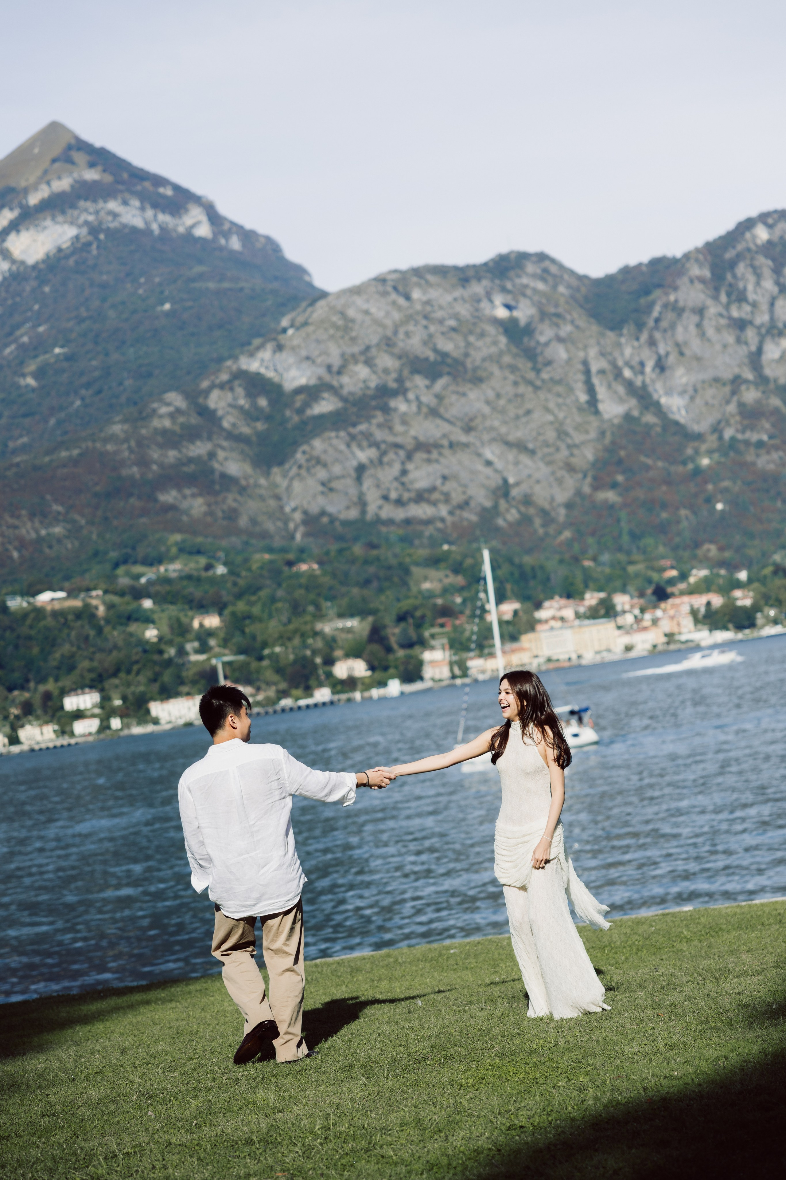 Couple holding hands beside Lake Como, with the dramatic mountains of Bellagio in the background.