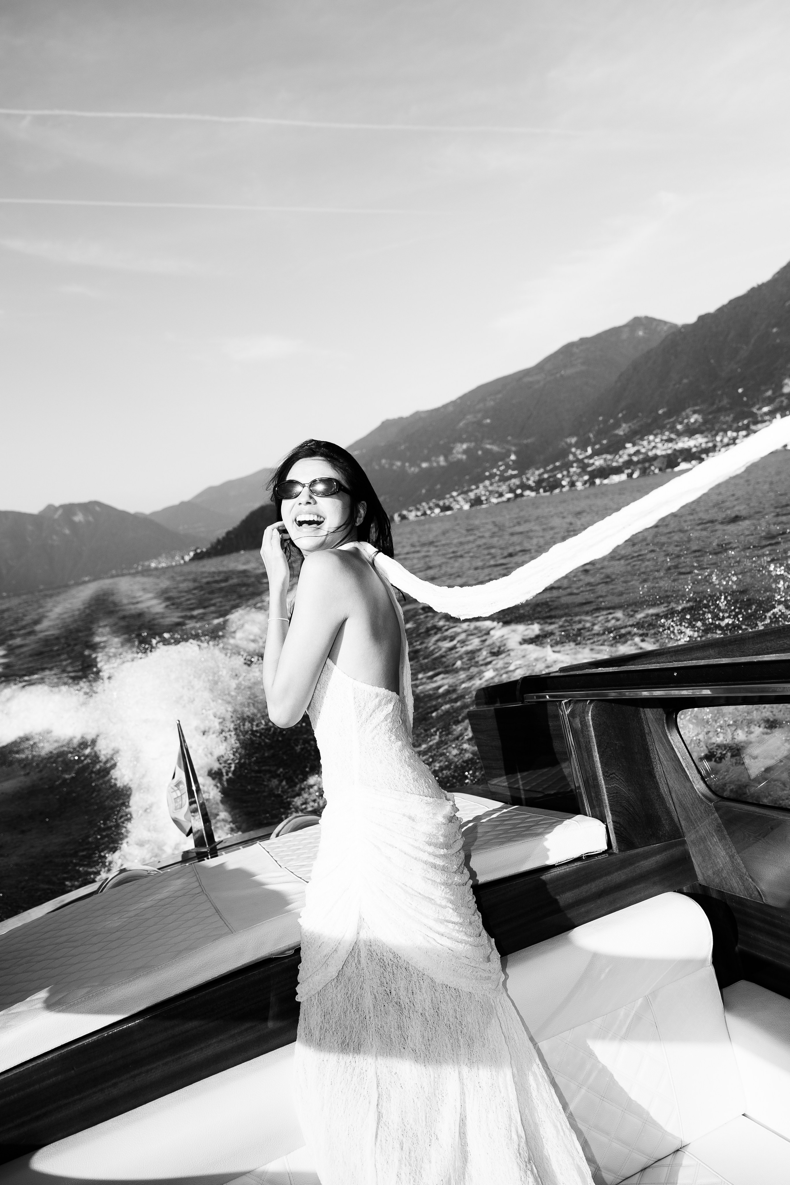 Black and white photograph of a smiling woman on a wooden boat with Lake Como and mountains behind her.