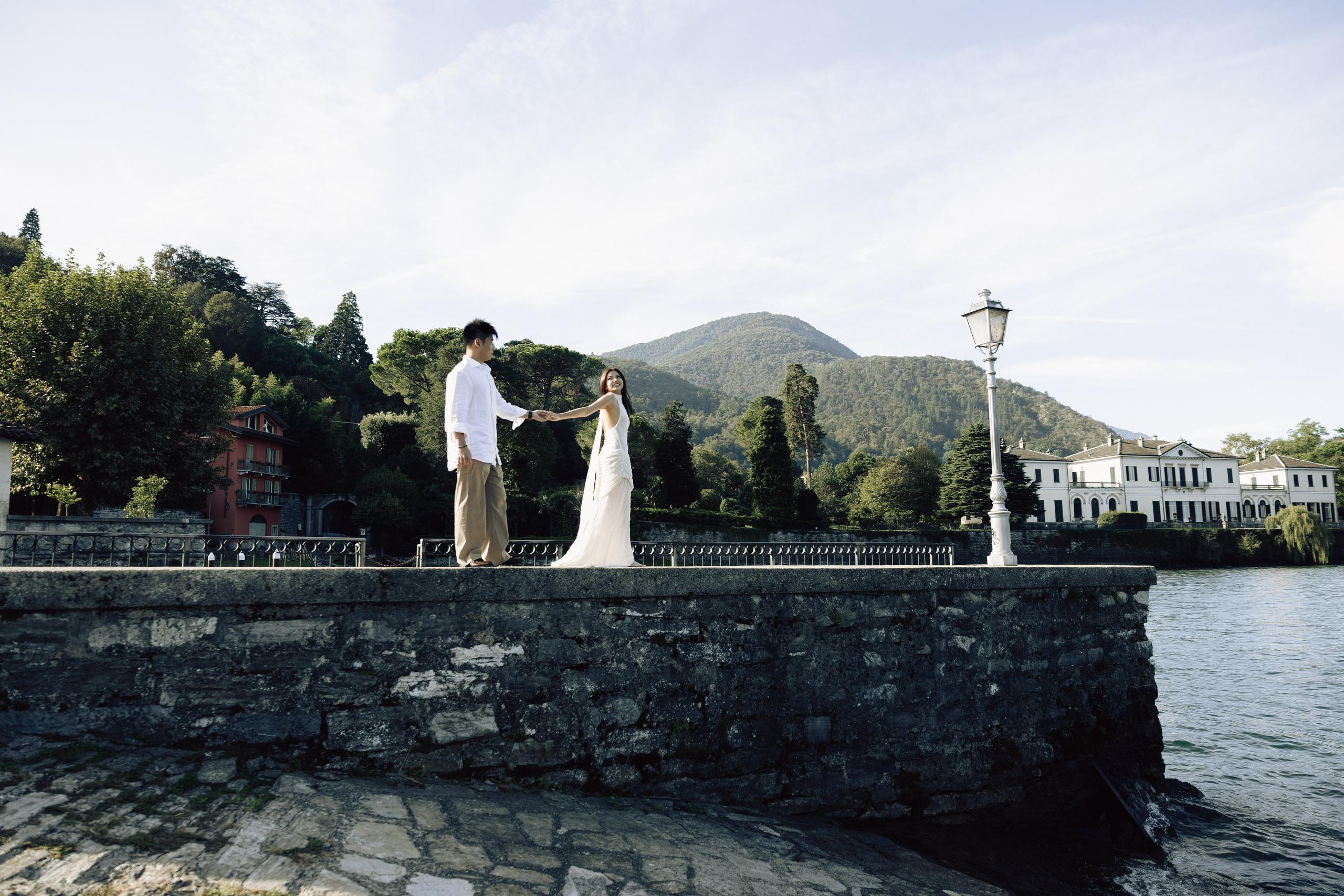 Couple holding hands on a stone waterfront terrace in Bellagio, with Lake Como and the surrounding mountains behind.