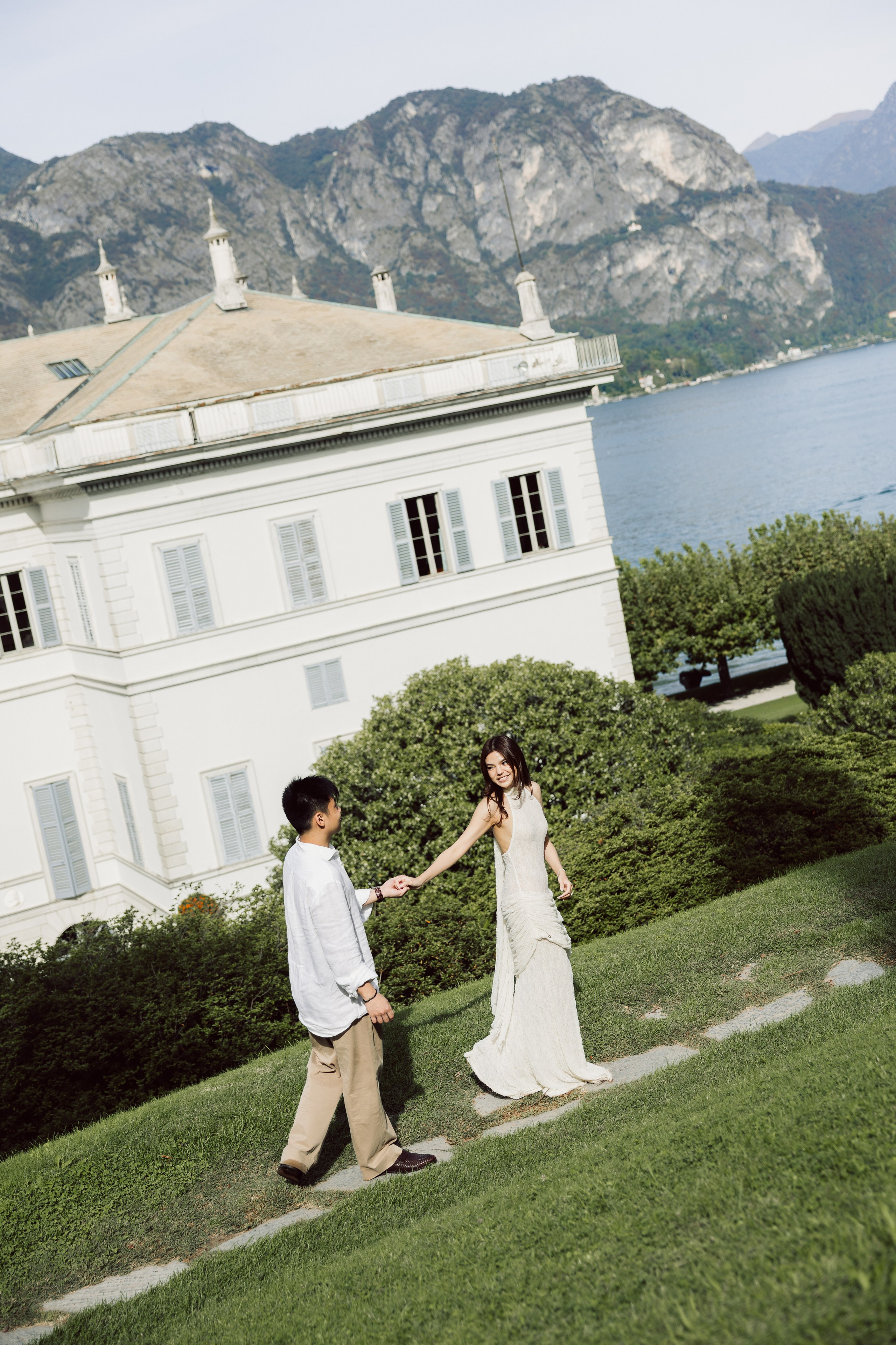 Couple holding hands while walking through the gardens of Villa Melzi in Bellagio, with the lake and mountains behind.